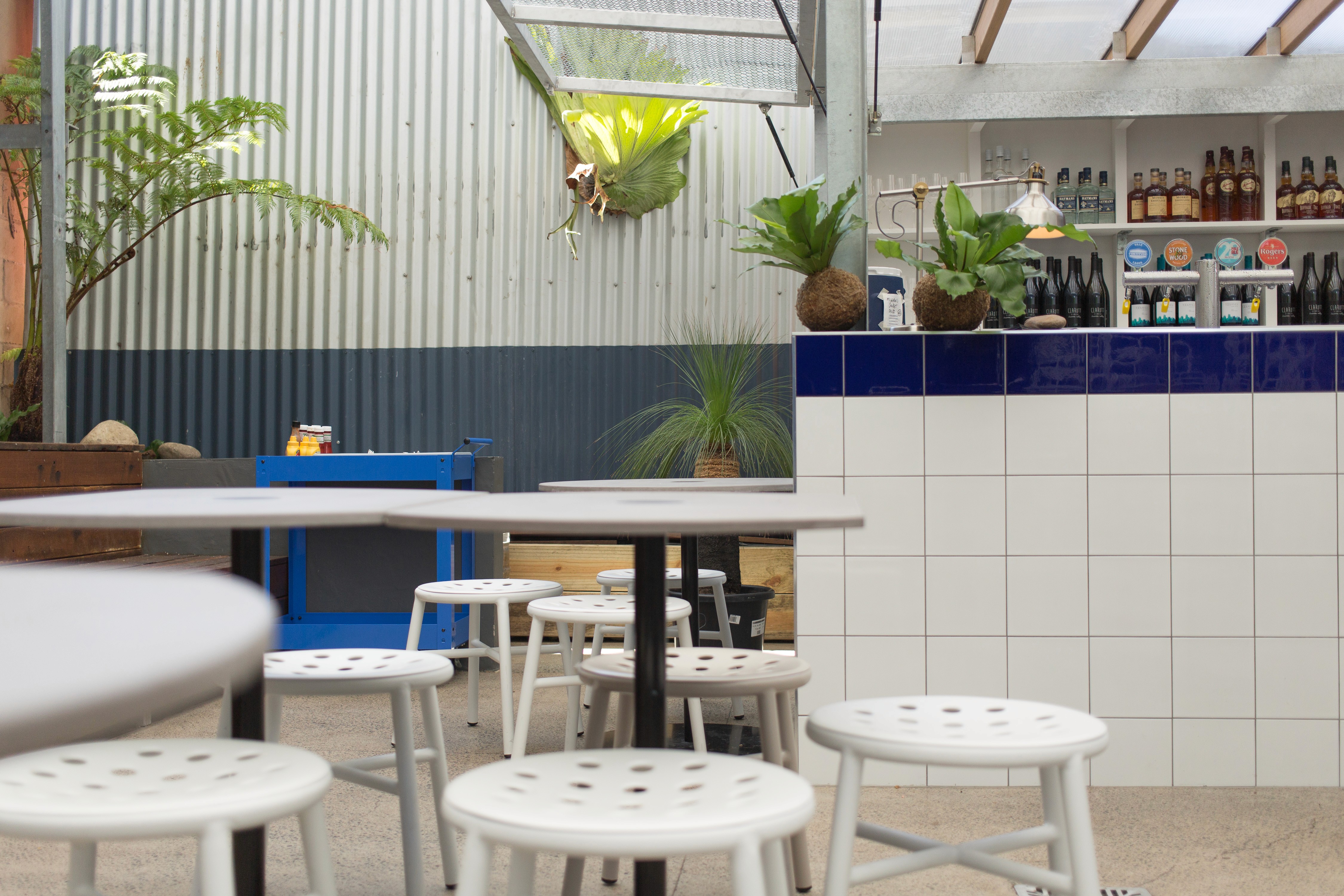 Courtyard seating and bar detail at Ben’s Burgers West End with white metal stools, blue-tiled counter, hanging plants, and corrugated metal backdrop.