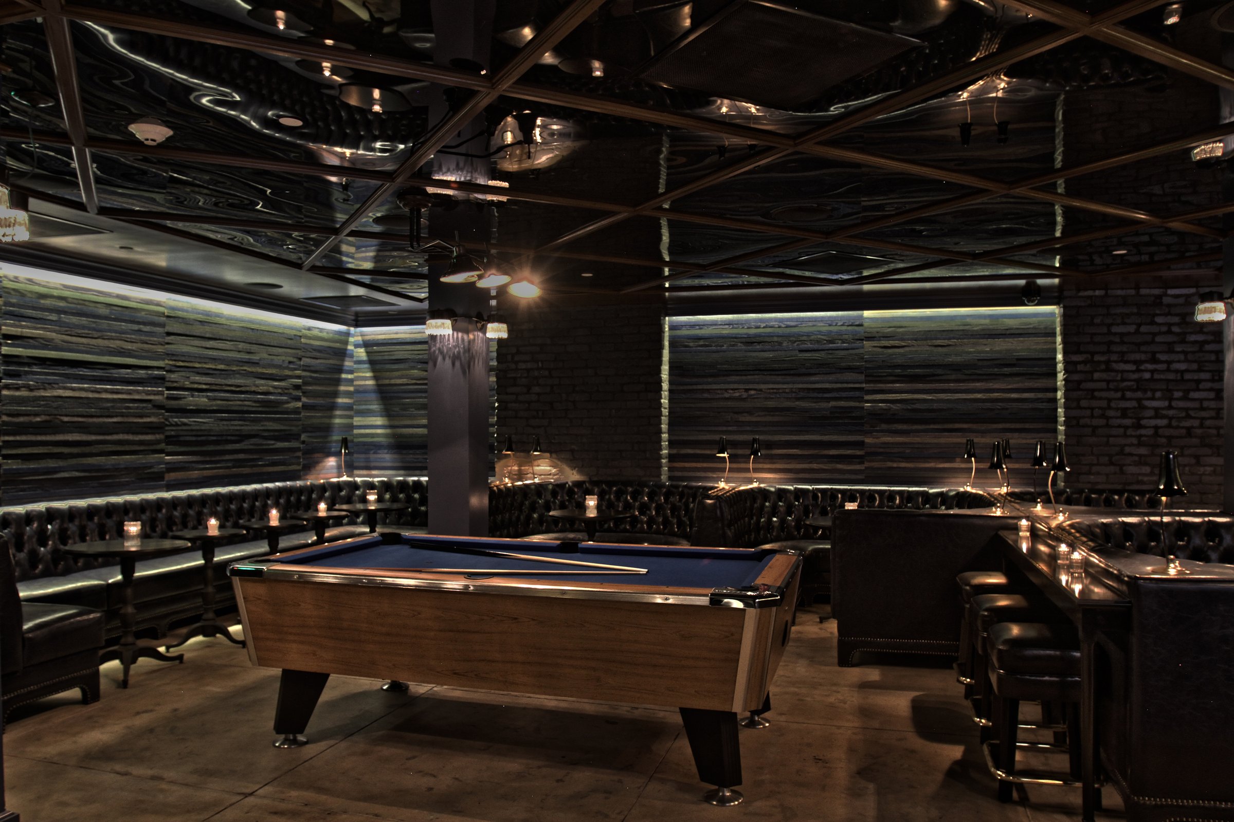 Pool table in a dim lounge with tufted perimeter seating and striped walls beneath an exposed ceiling.