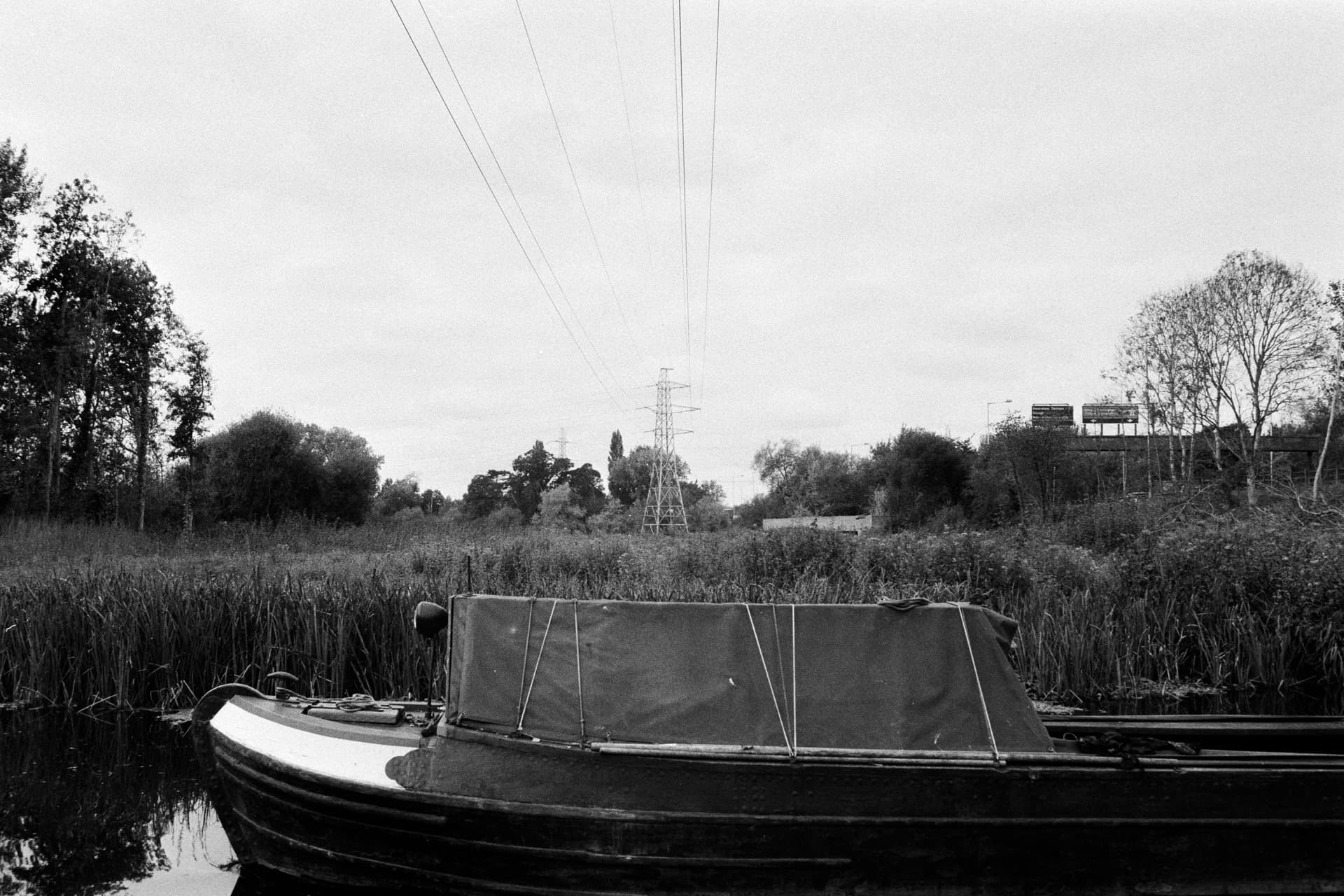 Moored narrowboat with canvas cover on overgrown canal with power lines overhead