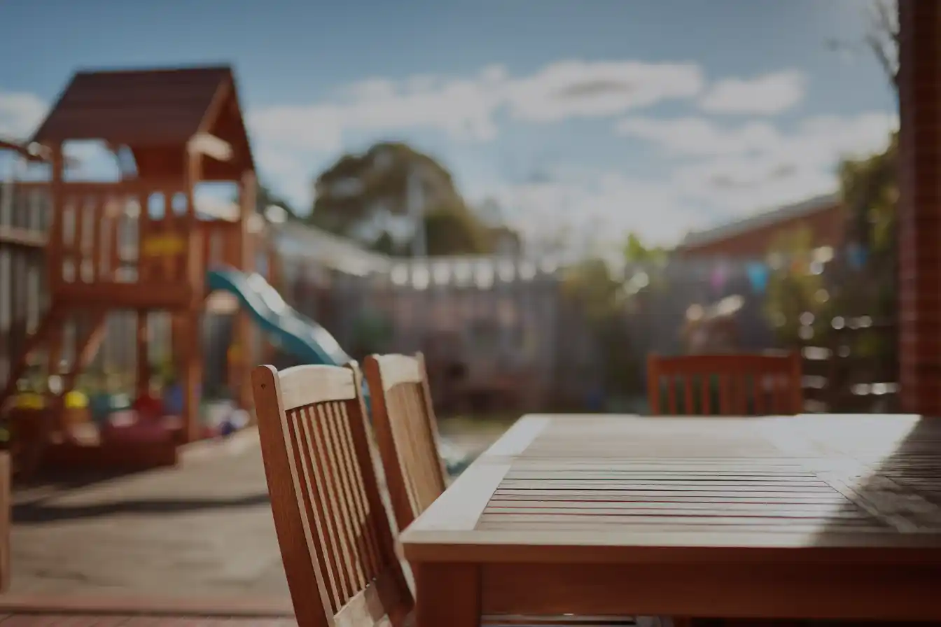 Wooden outdoor dining table with chairs in focus, set in a bright backyard. A blurry playground with a slide and a sunny sky in the background creates a warm, inviting atmosphere.