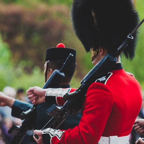 Twee soldaten in uniform, de een in een rode jas met een hoge zwarte hoed en de ander in een zwarte outfit met een rode pluim, met geweren in de hand.