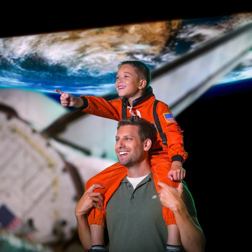 Ett barn i en orange dräkt sitter på en mans axlar, båda ler och pekar, med en bild av jorden från rymden i bakgrunden.