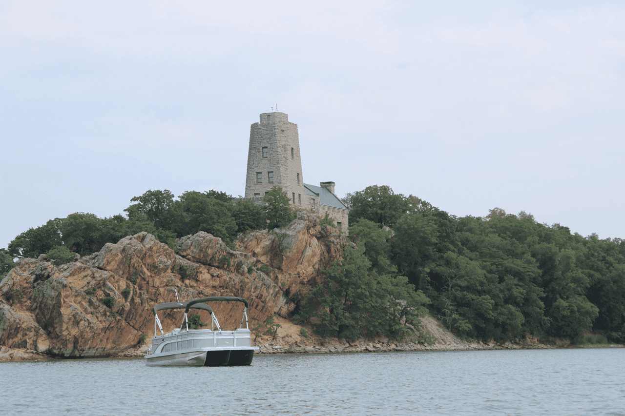 A motorboat cruises near a rocky shoreline topped with lush greenery and a unique, tower-like stone building under a cloudy sky.