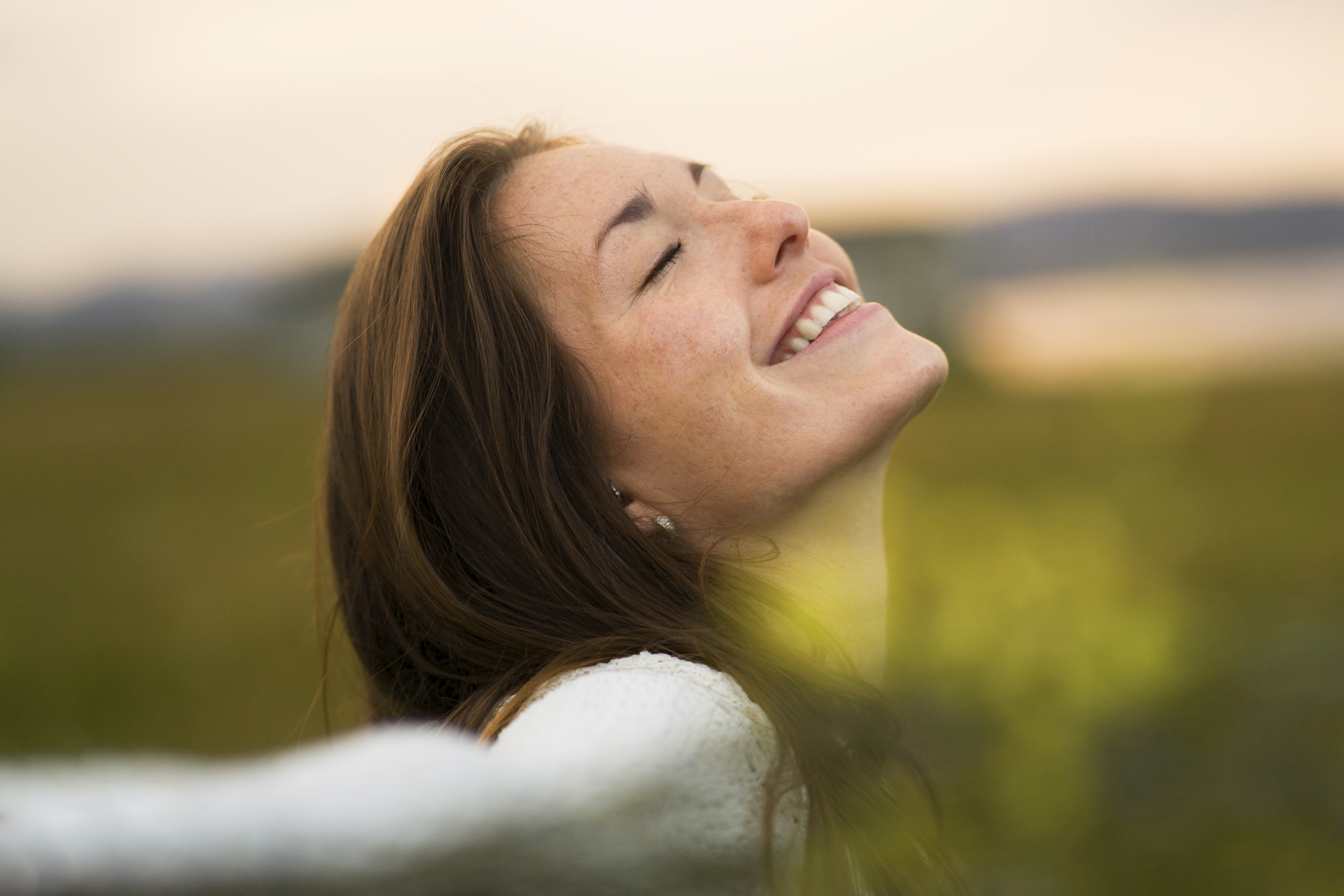 Close-up of a smiling woman with her eyes closed, tilted toward the sun in a peaceful outdoor setting.