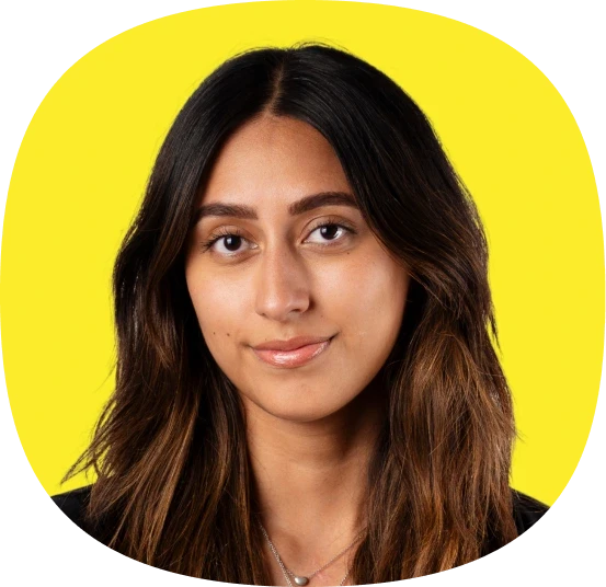A woman with long, dark hair and a slight smile poses in front of a bright yellow background. She is wearing a black top and a necklace.