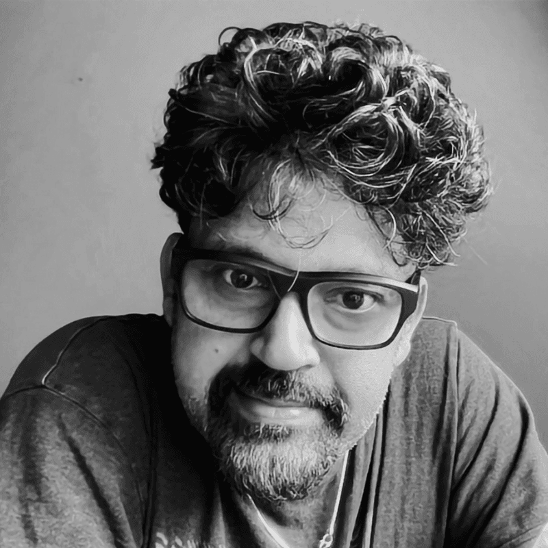An architect standing at a desk in a modern studio, reviewing architectural models and documents. Shelves filled with books and materials line the background, with natural light coming through large windows.