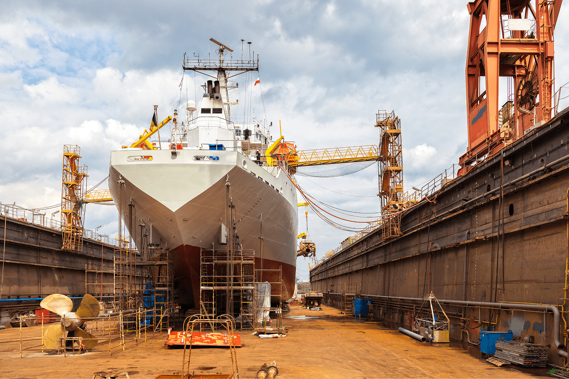 A ship is dry-docked in a shipyard, surrounded by structures and equipment under a cloudy sky.