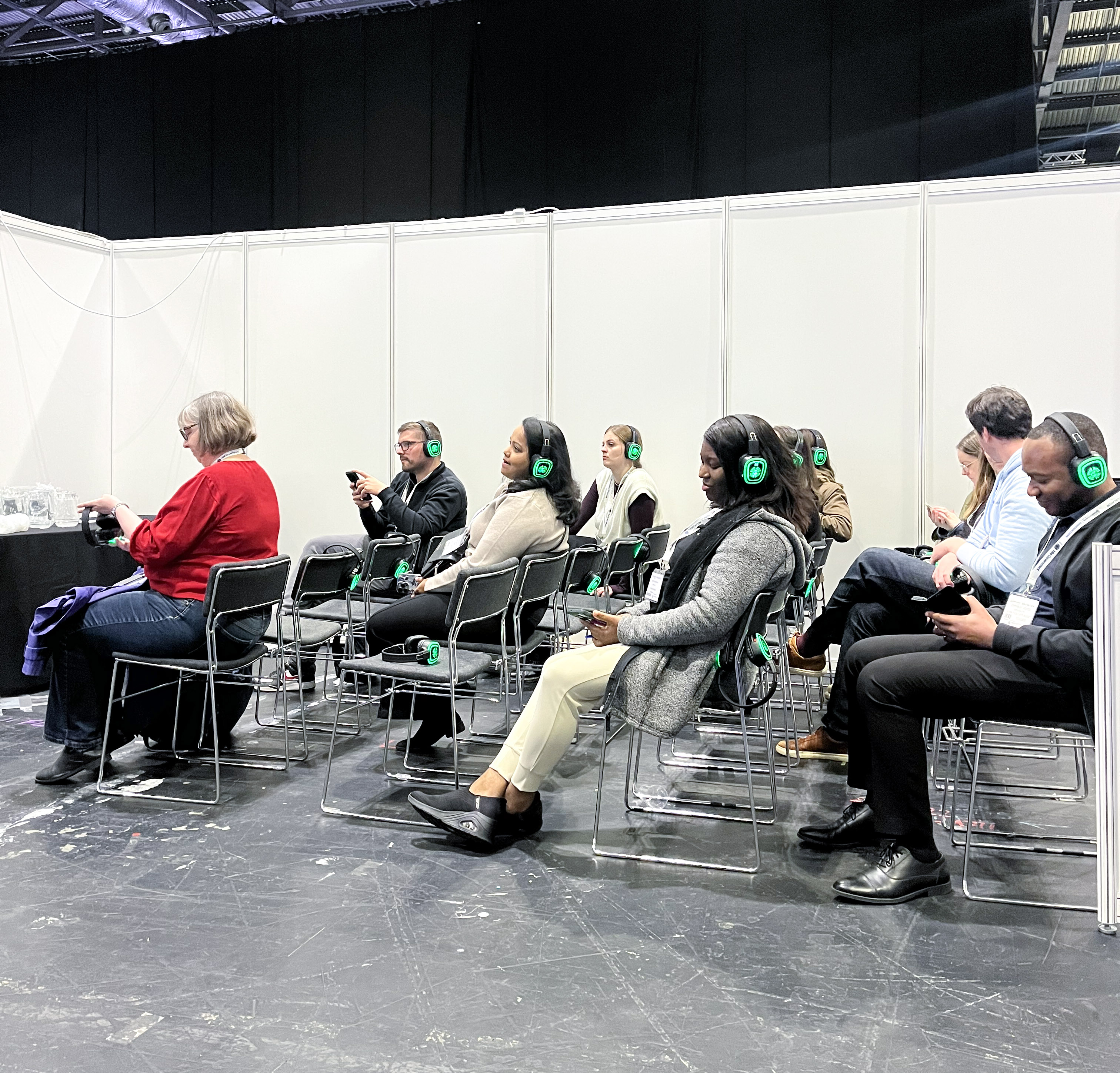 Audience members sit in rows of chairs at a conference session, all wearing green‑lit wireless headsets. Most attendees are looking down at their phones or listening quietly. The space is set within white partition walls, with a high industrial ceiling visible above. 
