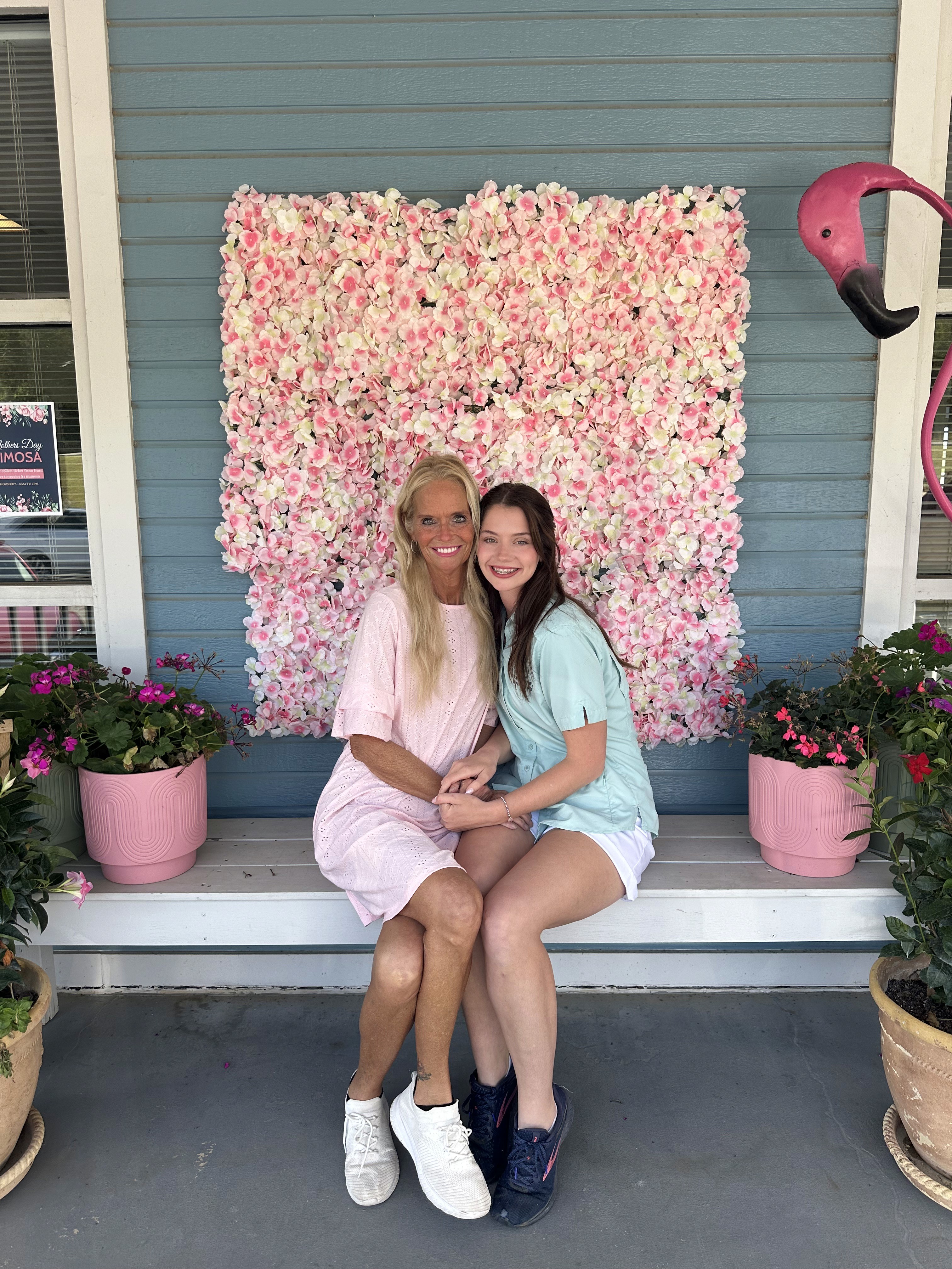 A mother and daughter sit on a bench in front of a decorative pink flower wall, with potted plants and a pink flamingo statue on either side.