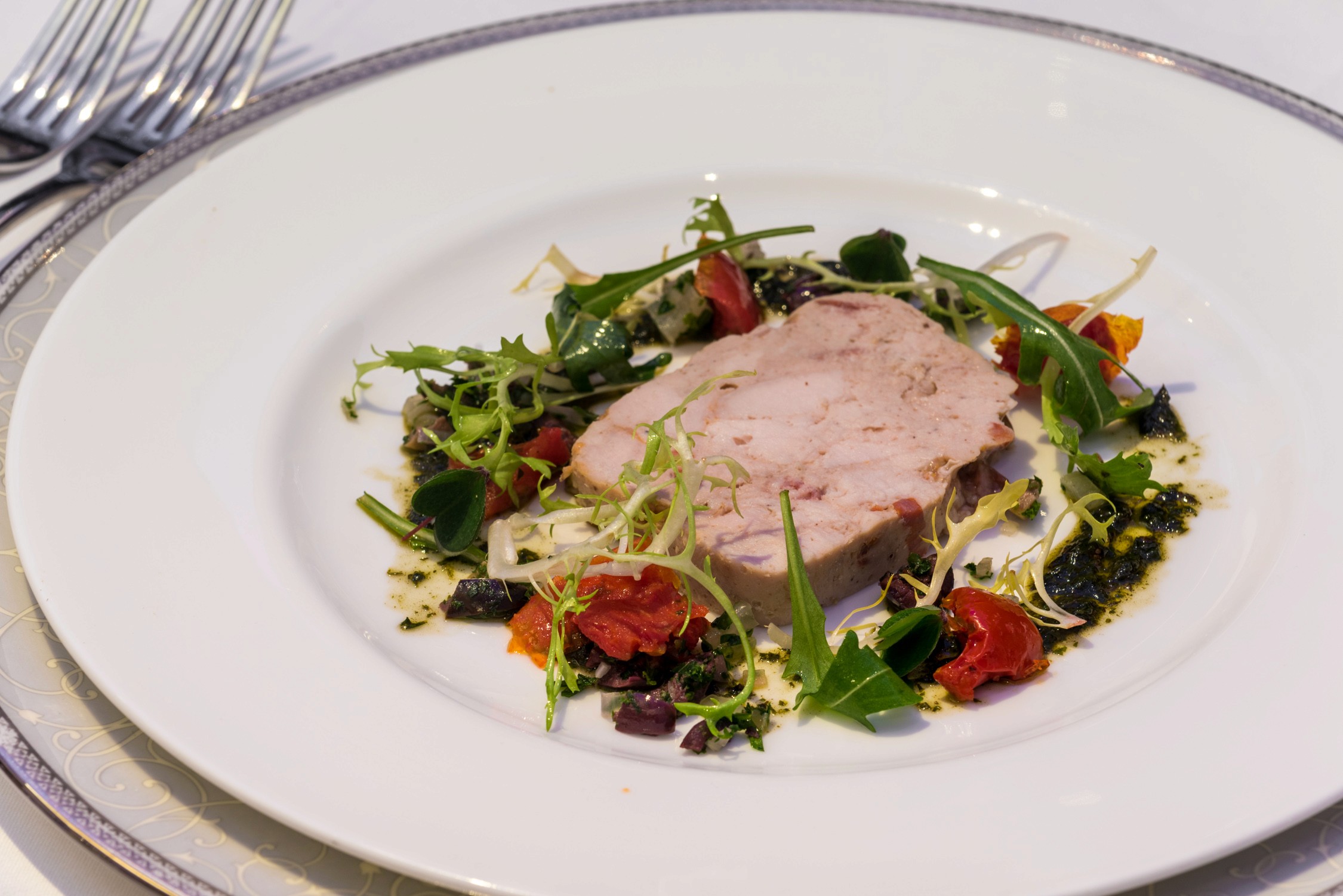 A plated dish featuring a slice of roasted meat topped with greens and cherry tomatoes on a white plate.