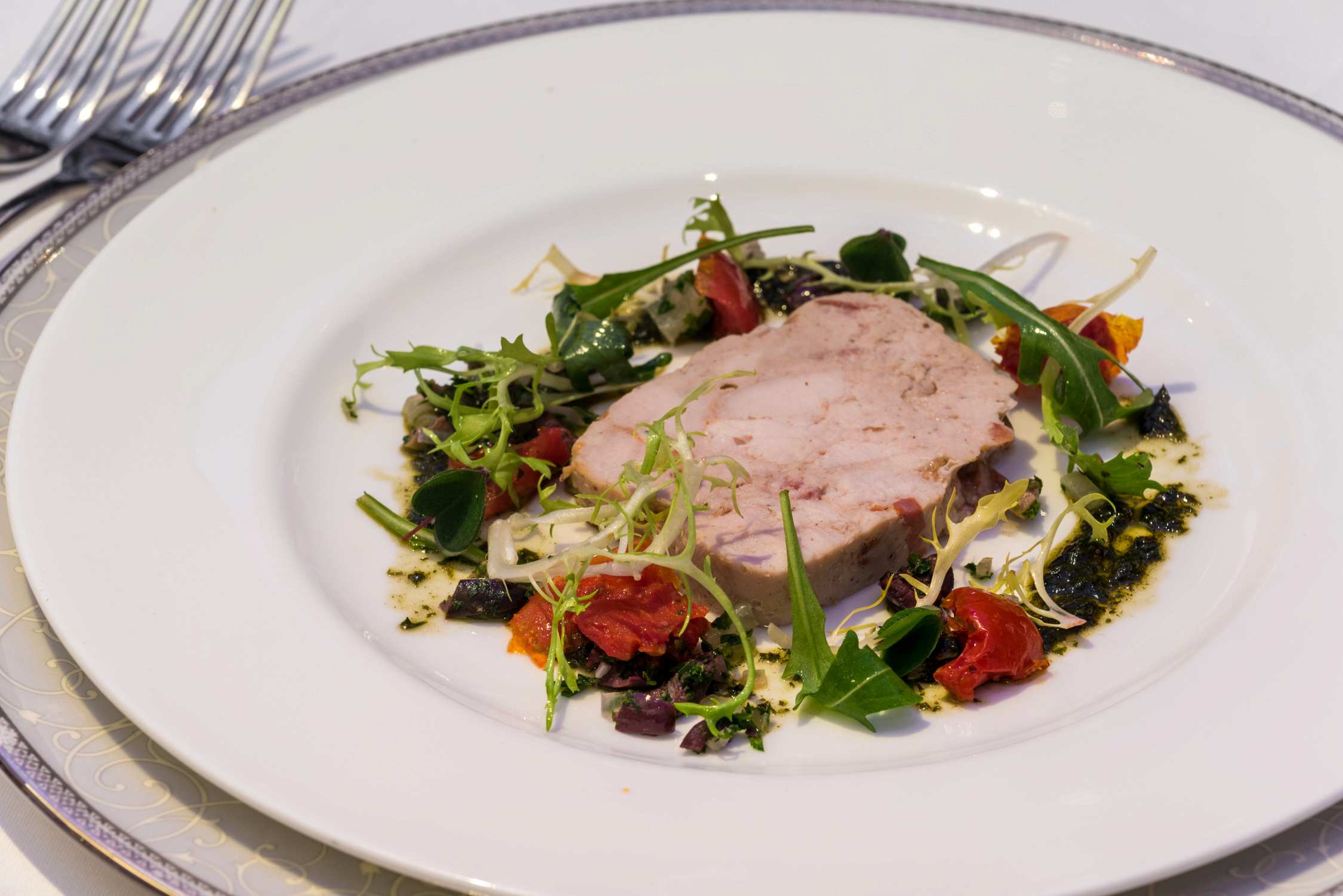 A plated dish featuring a slice of roasted meat topped with greens and cherry tomatoes on a white plate.