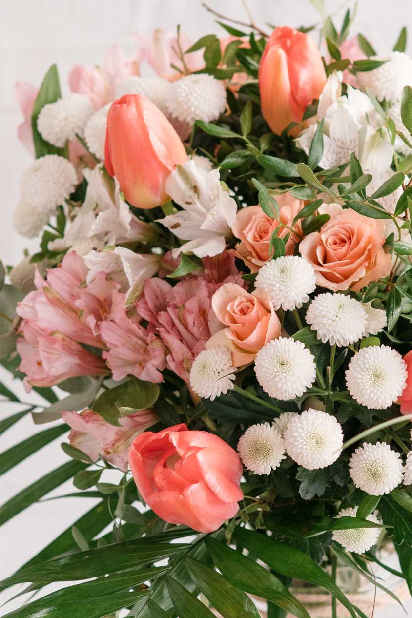 Primer pla d’un ram amb tulipes corall, roses préssec, crisantems blancs i alstromèries roses, envoltat de fulles verdes decoratives.