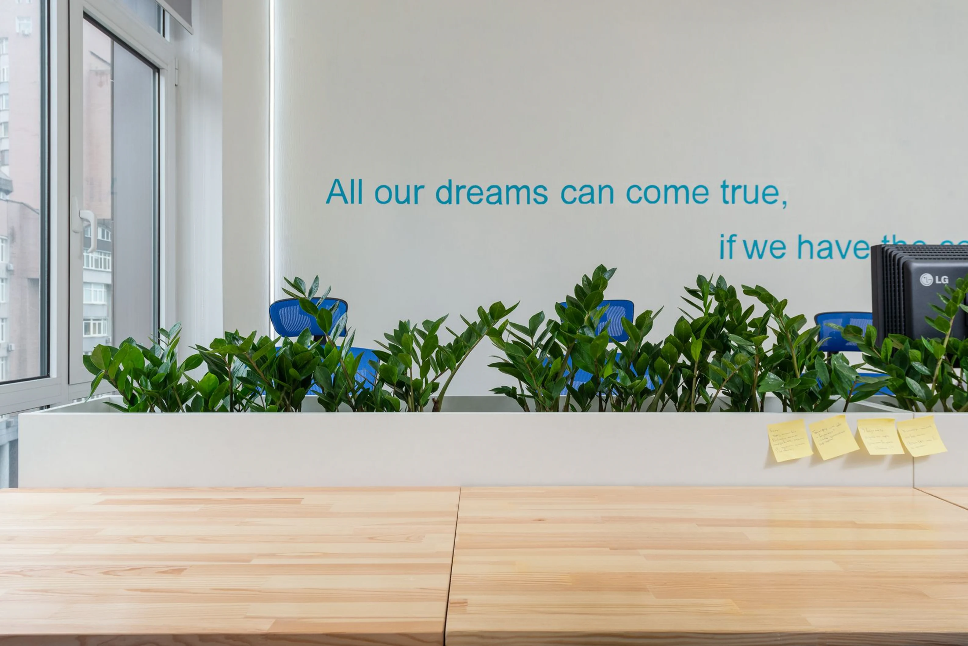 A bright, modern office workstation featuring a long wooden desk, a white planter box filled with green plants acting as a cubicle divider, and a blue inspirational quote painted on the background wall.