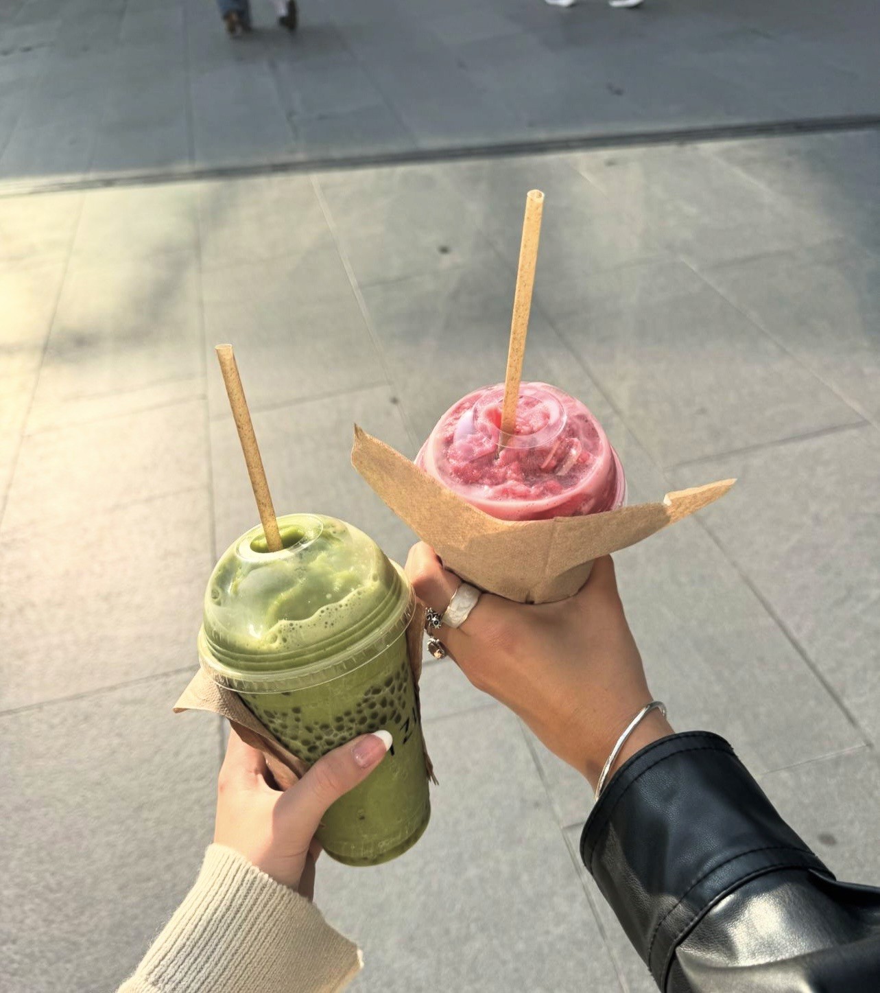 Two hands holding iced drinks with straws — one green matcha and one pink — outdoors on a overcast day.