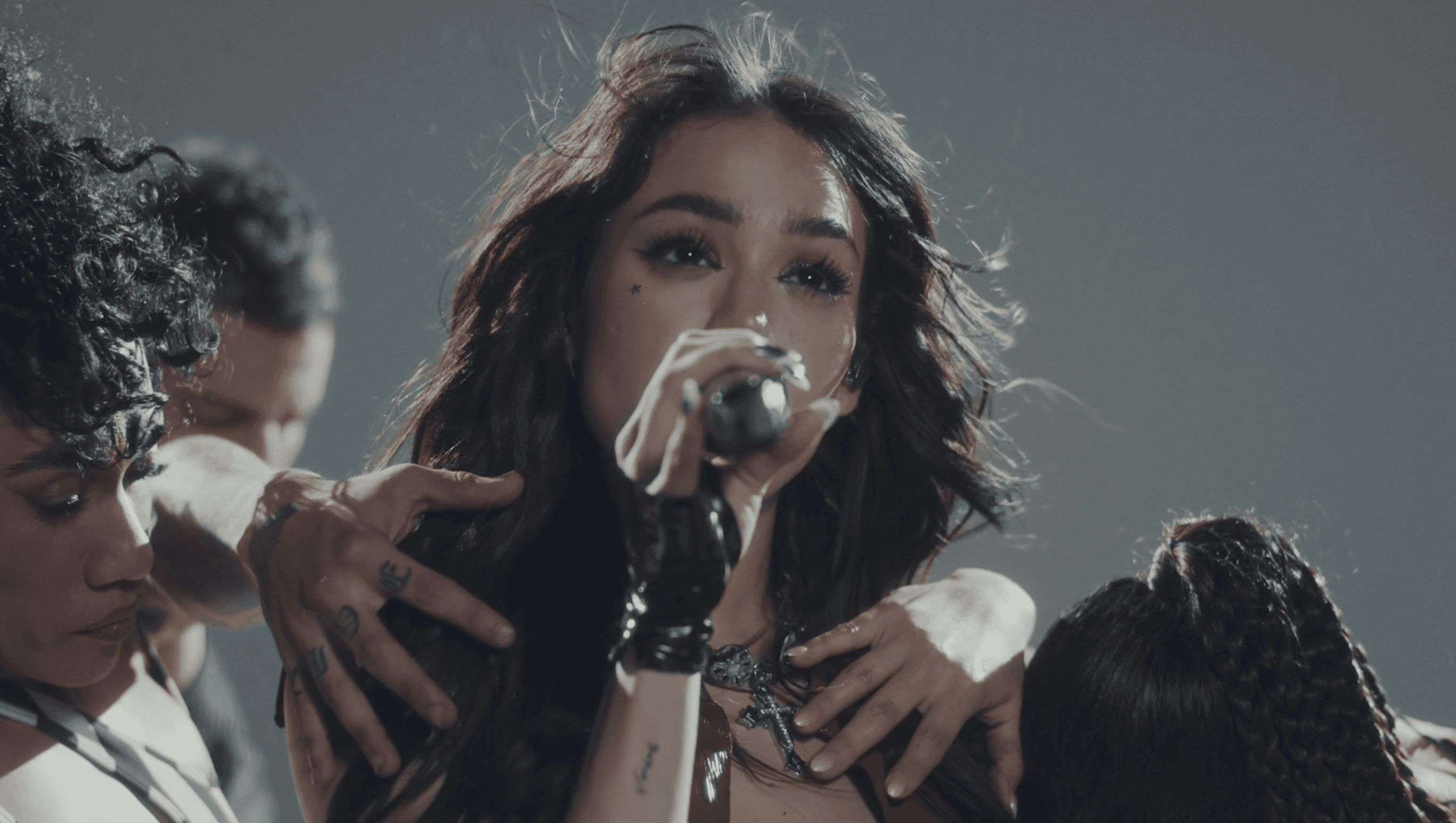 Black and white image of a Karol G smiling joyfully with closed eyes and long hair, wearing earrings and a sleeveless top. An MTV logo is in the corner.
