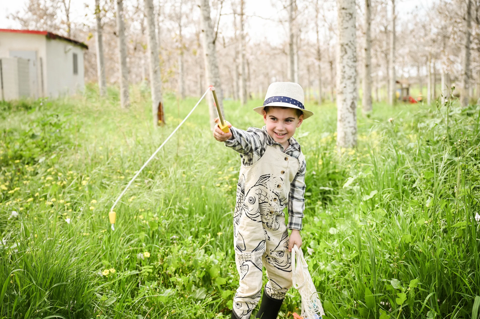 צילום קונספט יצירתי של ילד קטן ומתוק לבוש כדייג עם חכה קטנה וכובע קש, מחייך בלב חורשה ירוקה ופורחת. התמונה משדרת תמימות ושמחה של ילדות בטבע, מאת Shiriz Photography והצלמת שירי עציץ