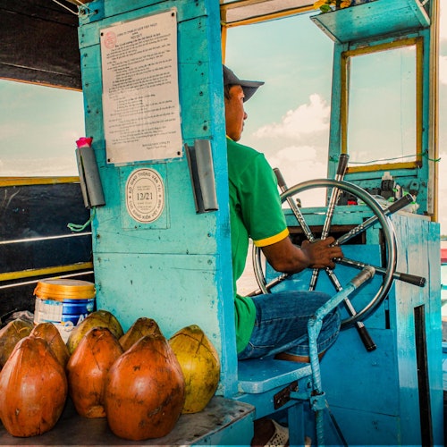 Seorang pria berbaju hijau mengemudikan perahu dengan kelapa dan wadah di sekitarnya, dengan latar langit dan air.