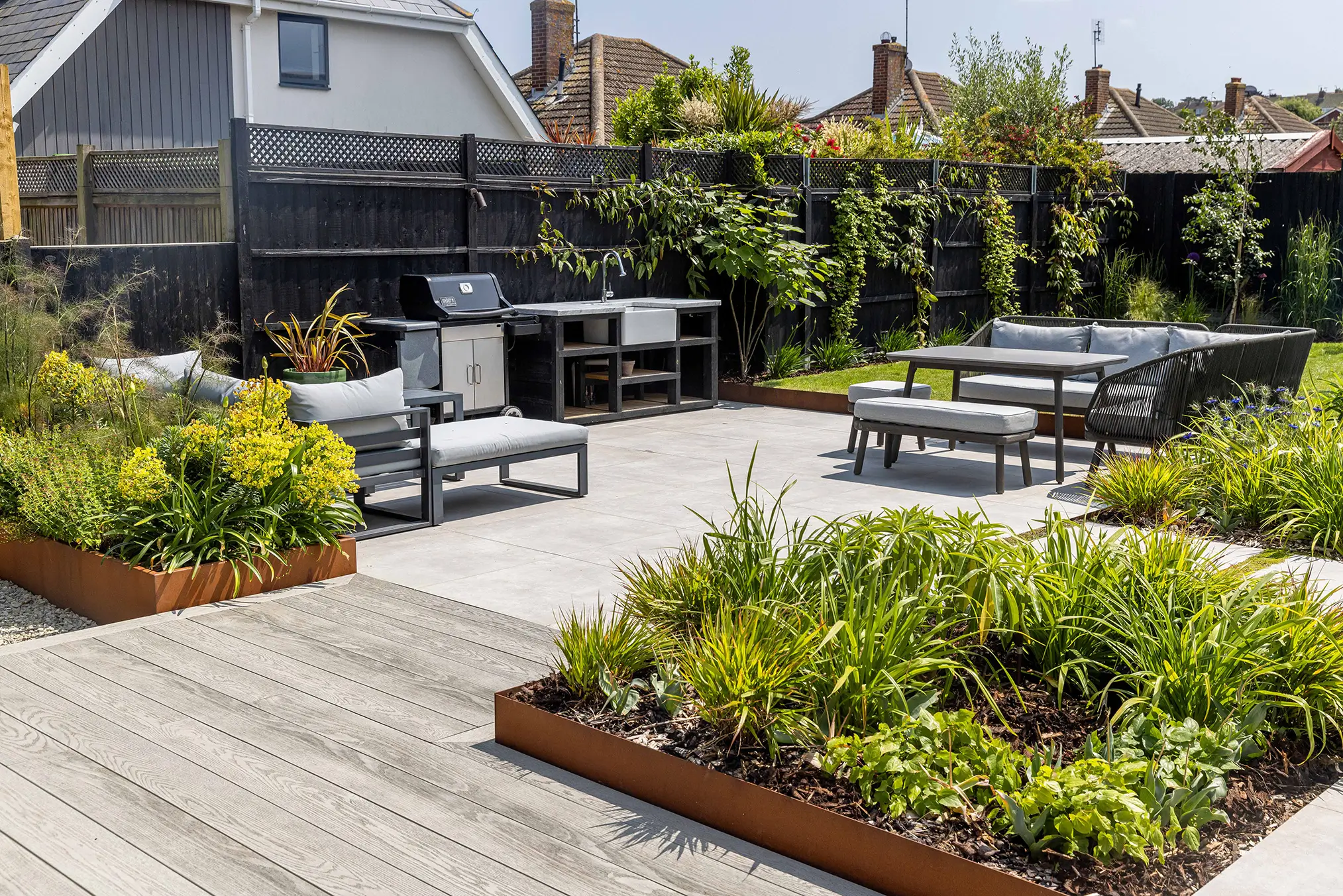 A sunny outdoor patio with wooden furniture, surrounded by lush greenery and garden beds.