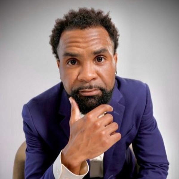 RJ Young with short curly hair and a beard, wearing a dark blue suit and white shirt, sits with his hand on his chin, looking thoughtfully at the camera against a plain background.