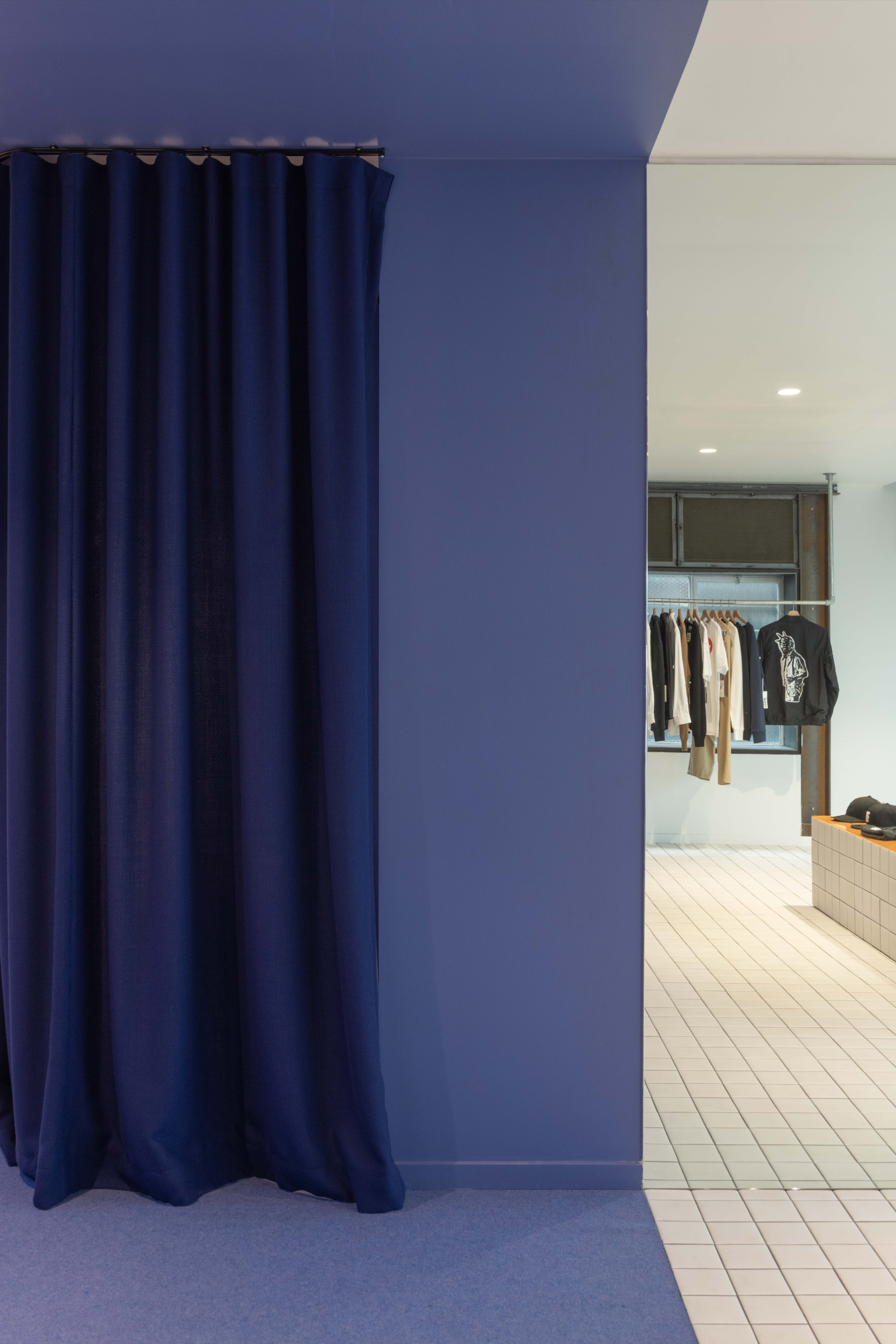 Deep blue curtained threshold inside the 199A® space, framing a transition from soft carpeted gallery area into a bright tiled retail zone with clothing rails and display plinths beyond.