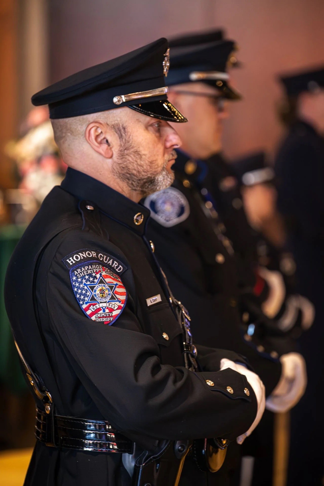 Three uniformed honor guards stand solemnly in a line, their detailed black uniforms adorned with badges, buttons, and an American flag patch, reflecting a formal ceremony setting.