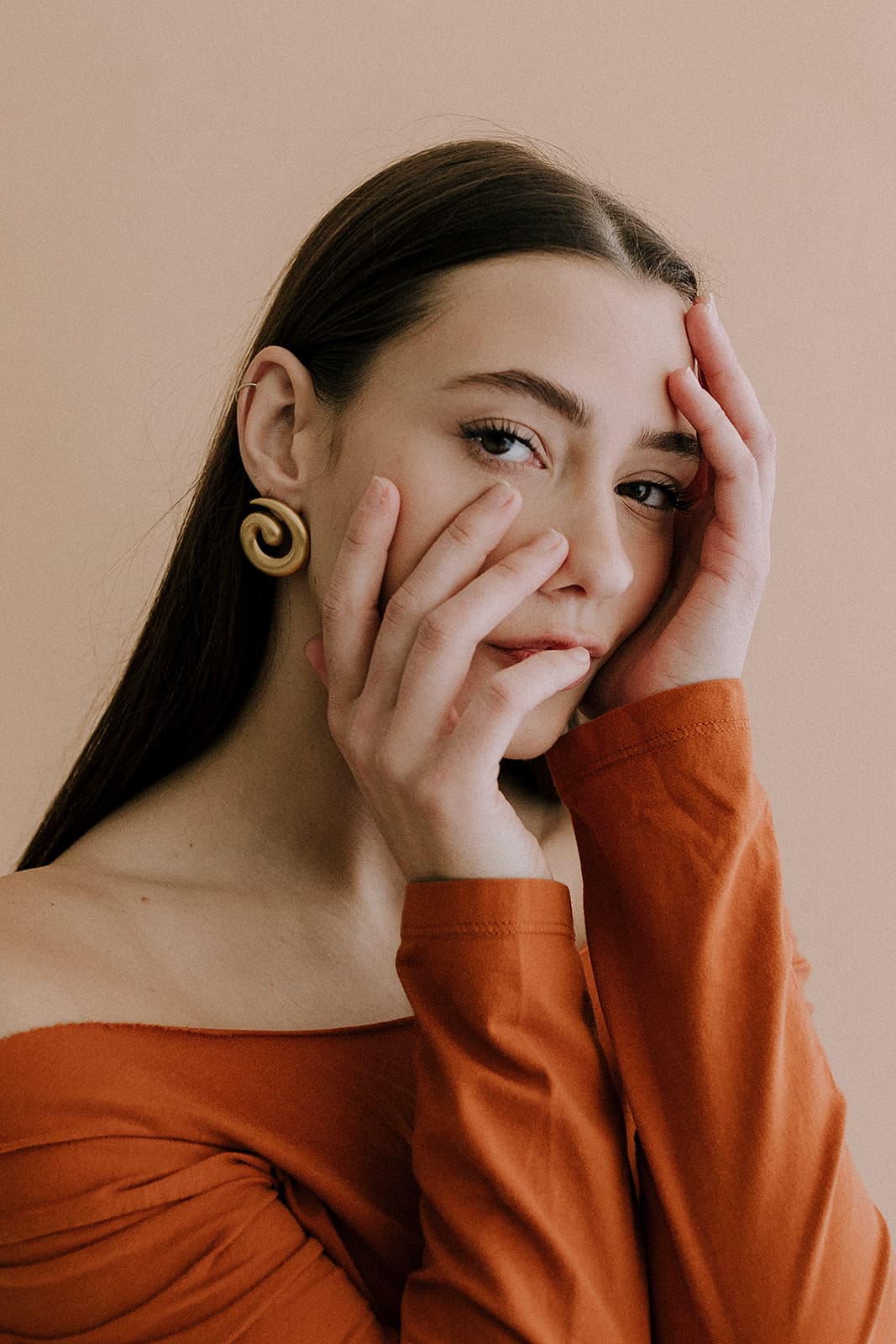 A close-up portrait of a model in an orange off-shoulder top, framed by her hands, taken in the natural light at Revelator Studio in Shreveport.