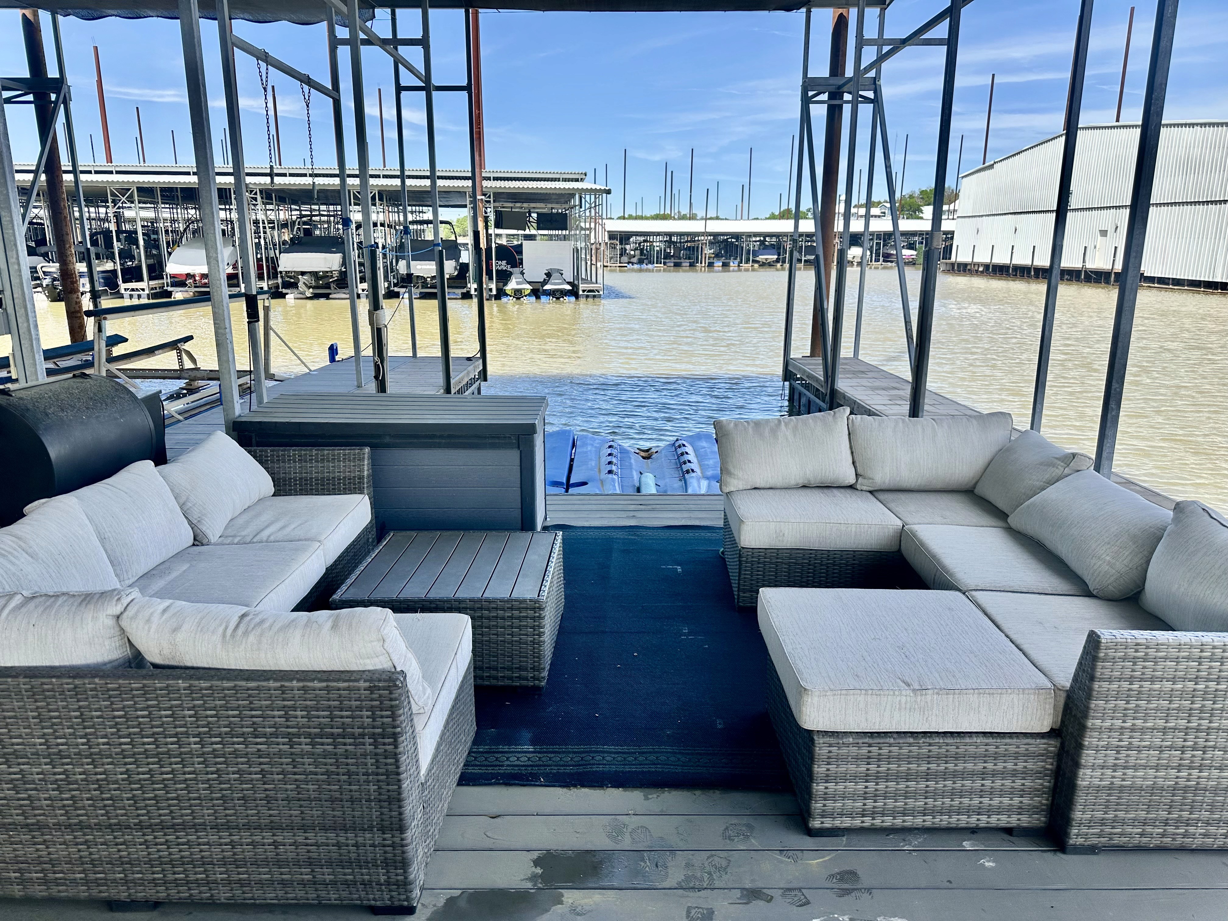 Outdoor patio with a wicker sofa set and cushioned seating on a dock overlooking a marina with calm water and boats under a clear blue sky.