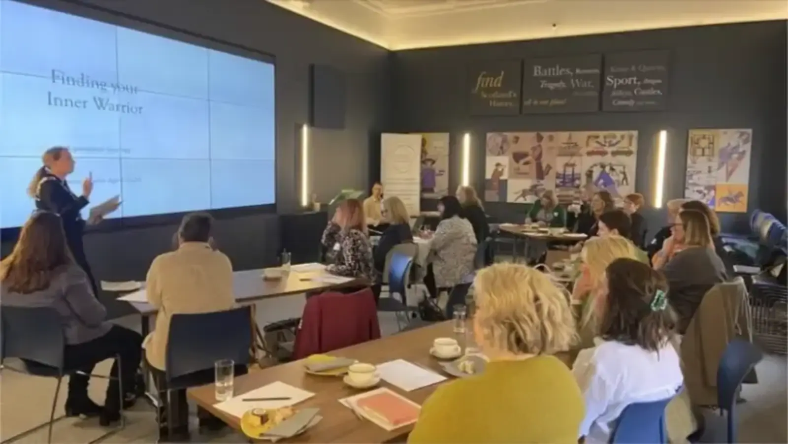 A speaker presents a "Finding Your Inner Warrior" slide to an engaged audience in a well-lit conference room. Attendees sit at tables, taking notes.