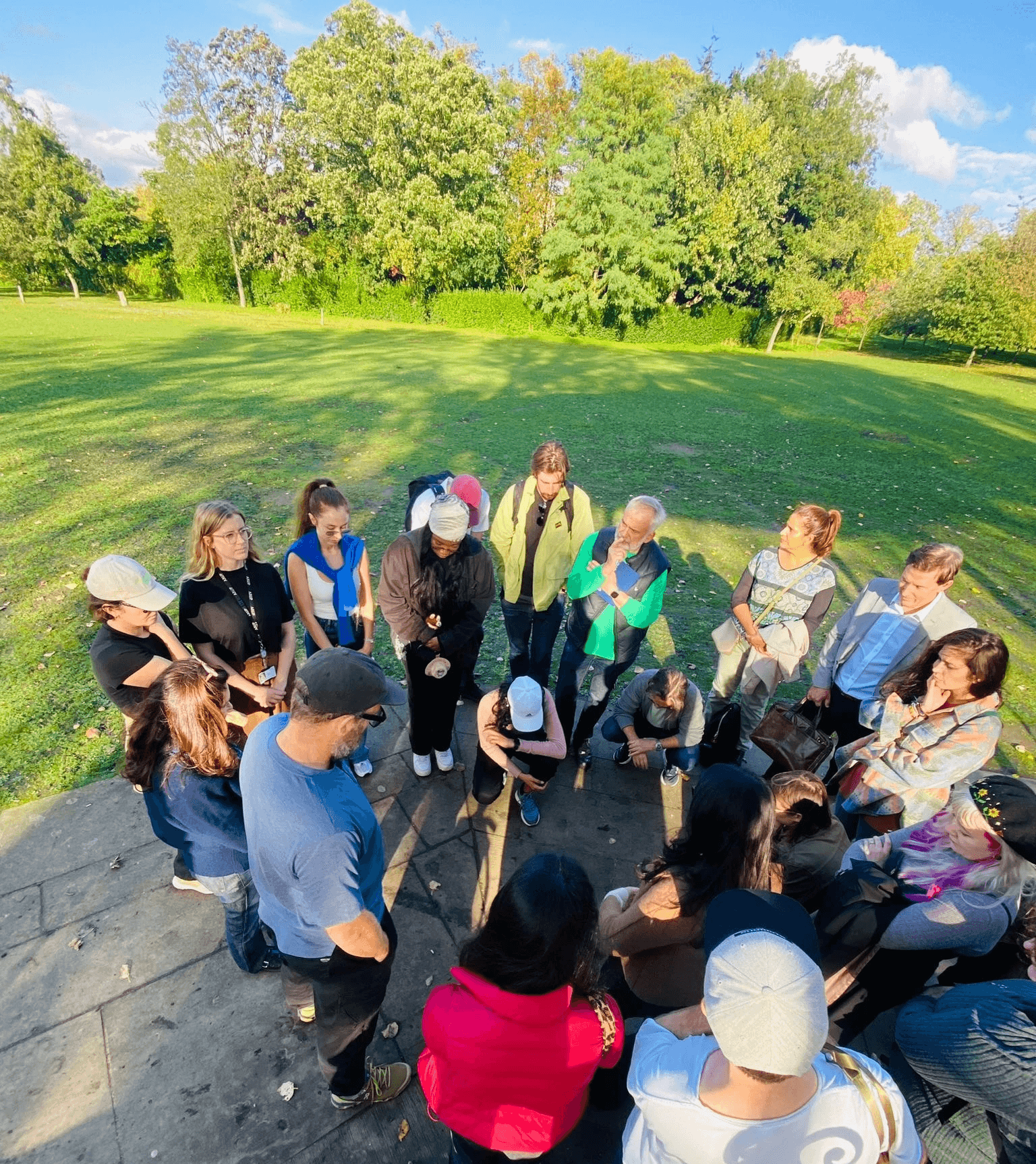 People gathered in a circle, listening to an audio message representation of non-human persona in Regent's Park, London, on a sunny day