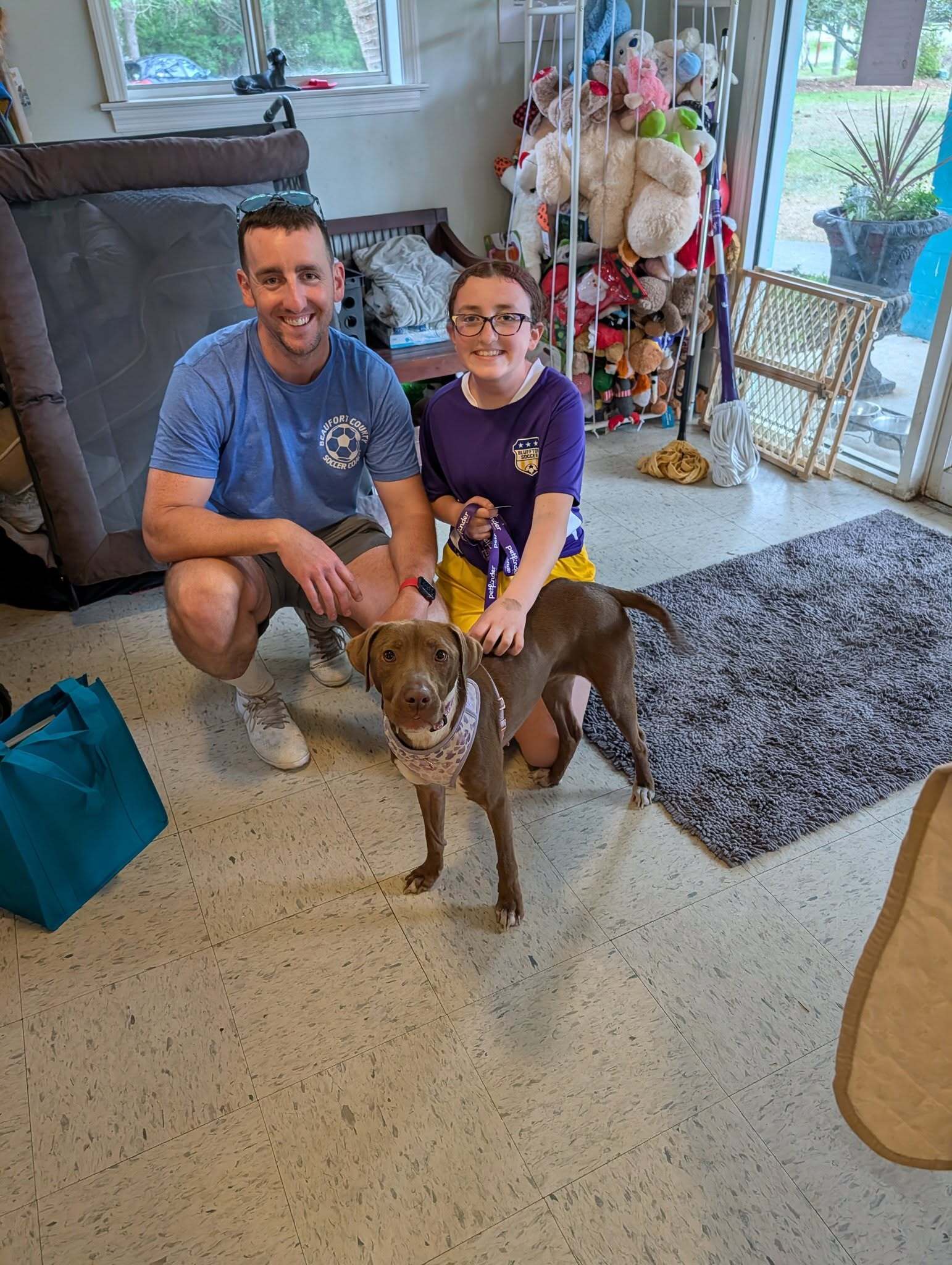 A man and a child smile while kneeling next to a dog in a cozy indoor space.