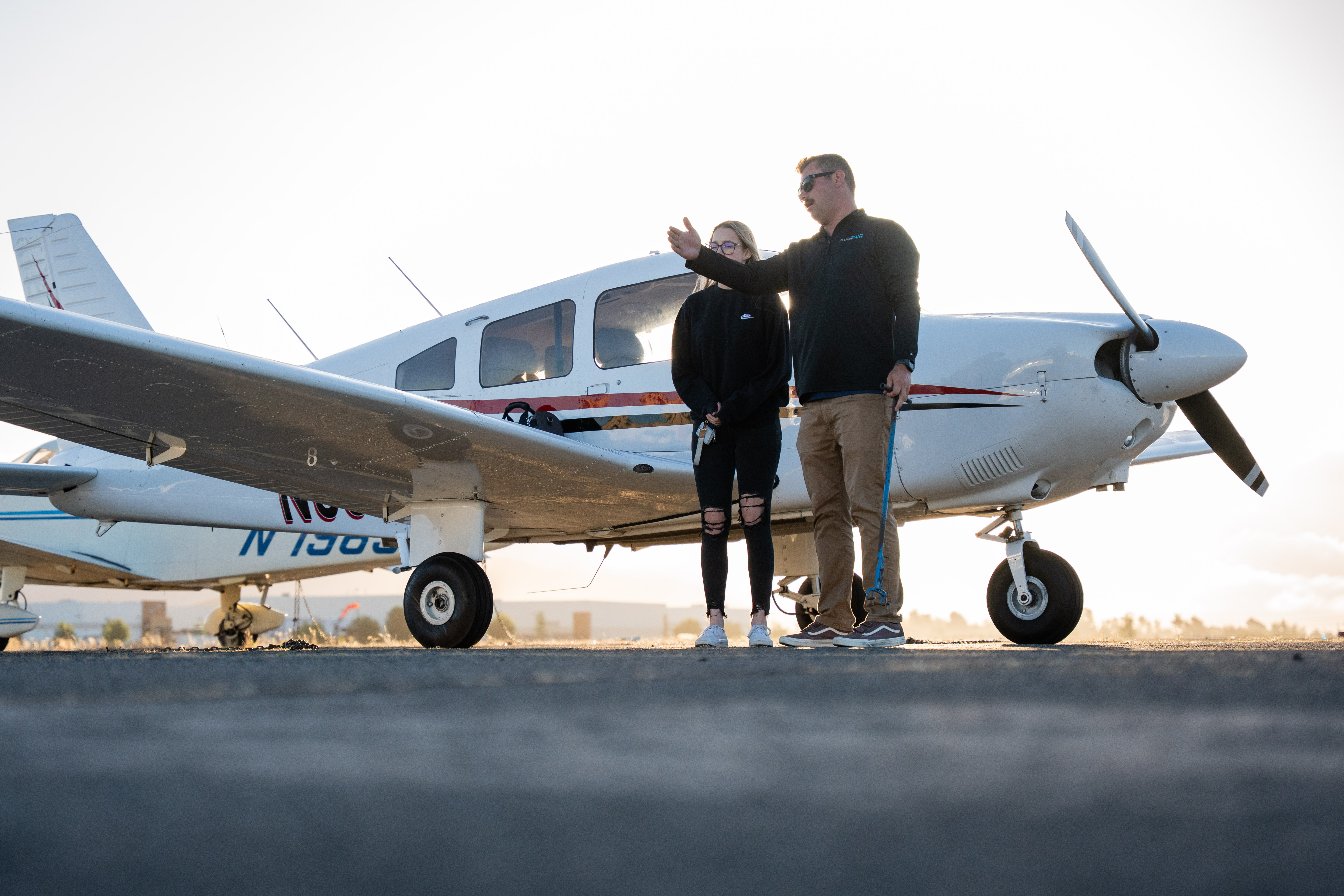 Modern open office lounge with large arched windows, plants, and soft round seating in white and terracotta tones, with people walking and working on laptops.