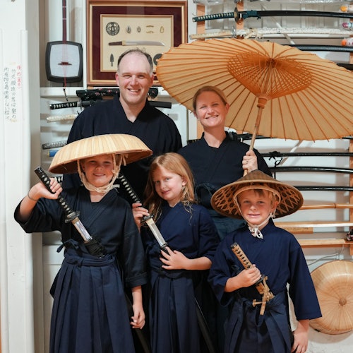 Een familie gekleed in traditionele Japanse kleding, met zwaarden en paraplu's in de hand, staat voor een tentoonstelling van zwaarden.