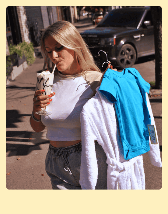 дівчина, світле волосся, пʼє молочний коктейль на вулиці і тримає одяг