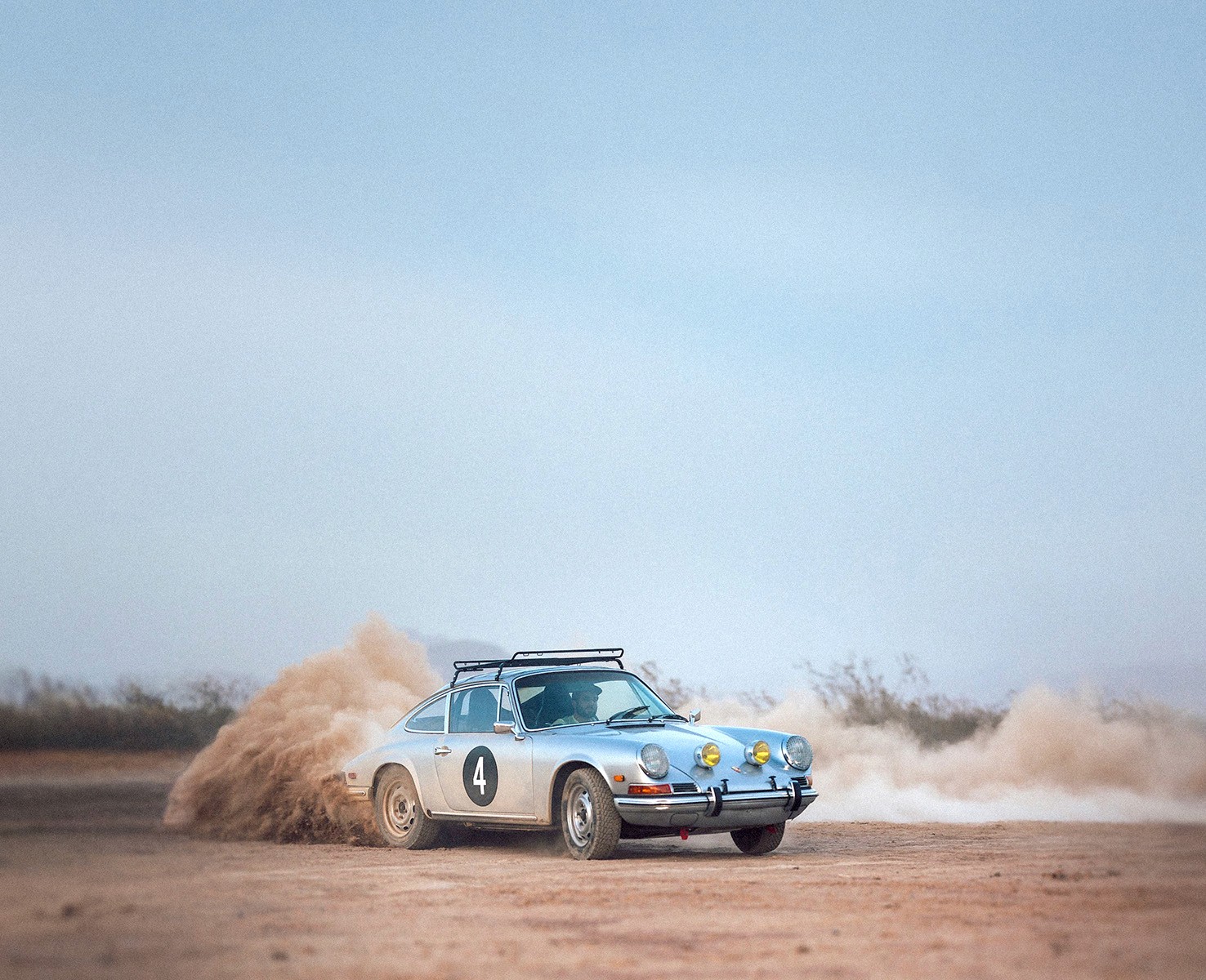 A vintage silver Porsche rally car with the number 4 on its side kicks up a cloud of dust as it drifts on a dirt track in a desert landscape.