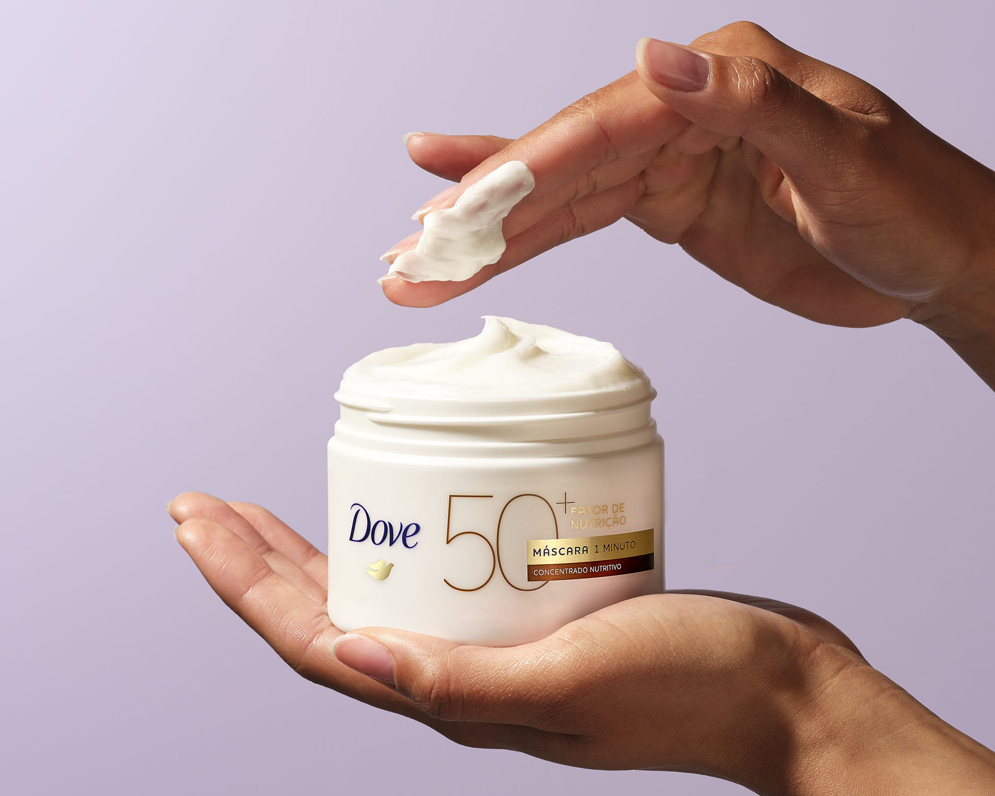 A person’s hand is scooping cream from a jar, which is held in the other hand, against a soft purple background.