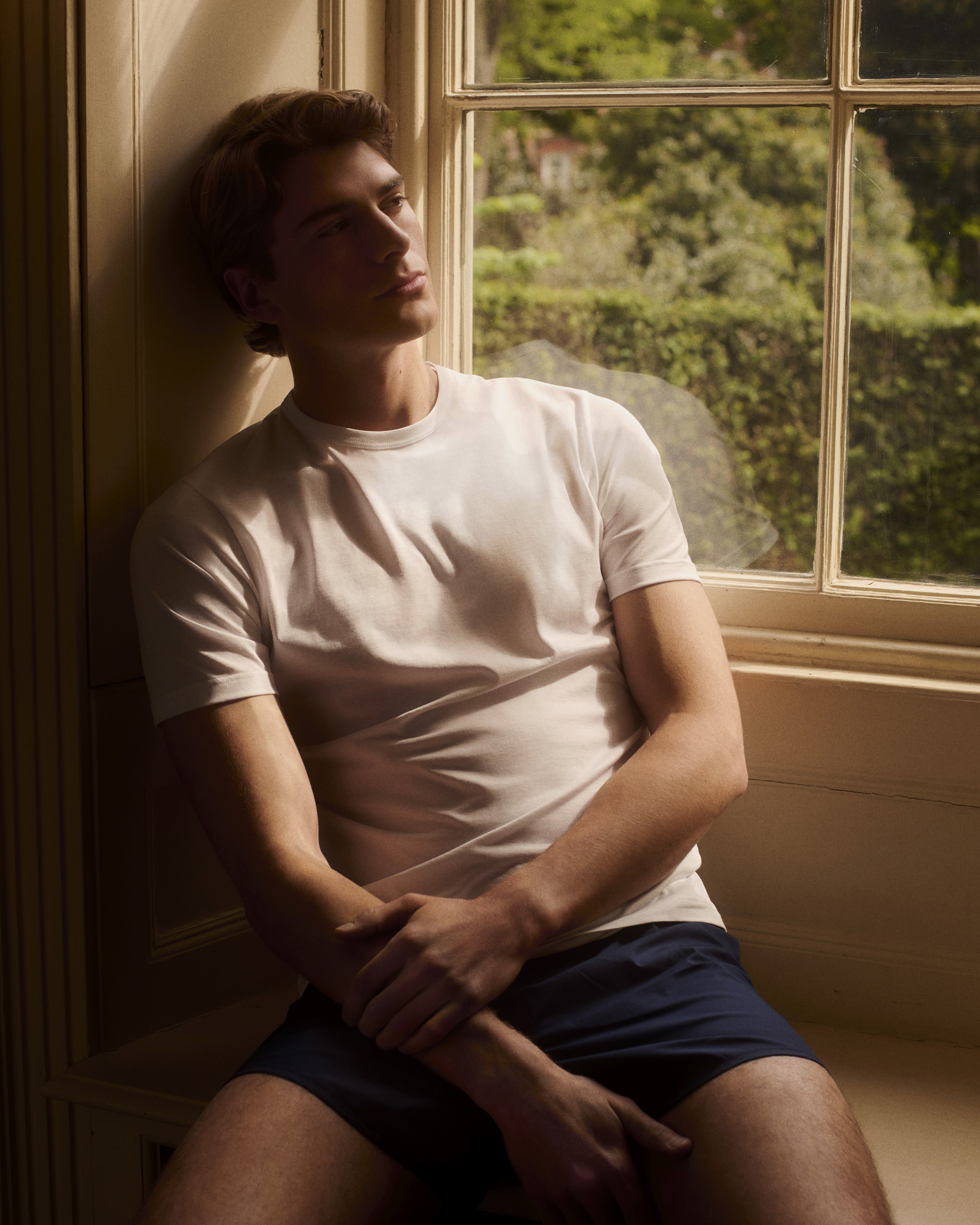 Dans le lookbook Sunspel, un homme porte un t-shirt blanc et un boxer devant une fenêtre