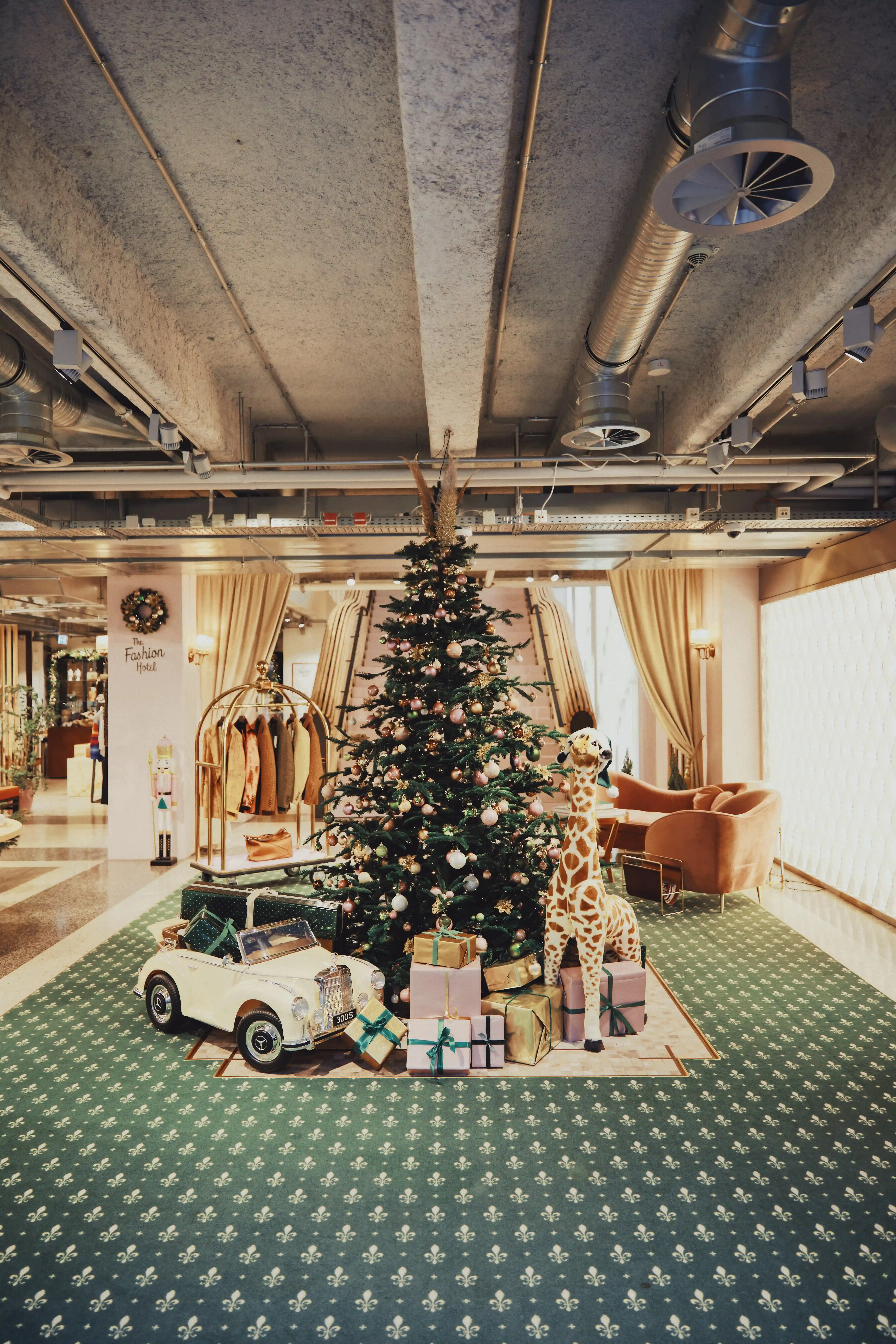 Large decorated Christmas tree at UZWEI on a green patterned carpet, surrounded by a plush giraffe, a vintage toy car, wrapped presents and a golden clothes rack.