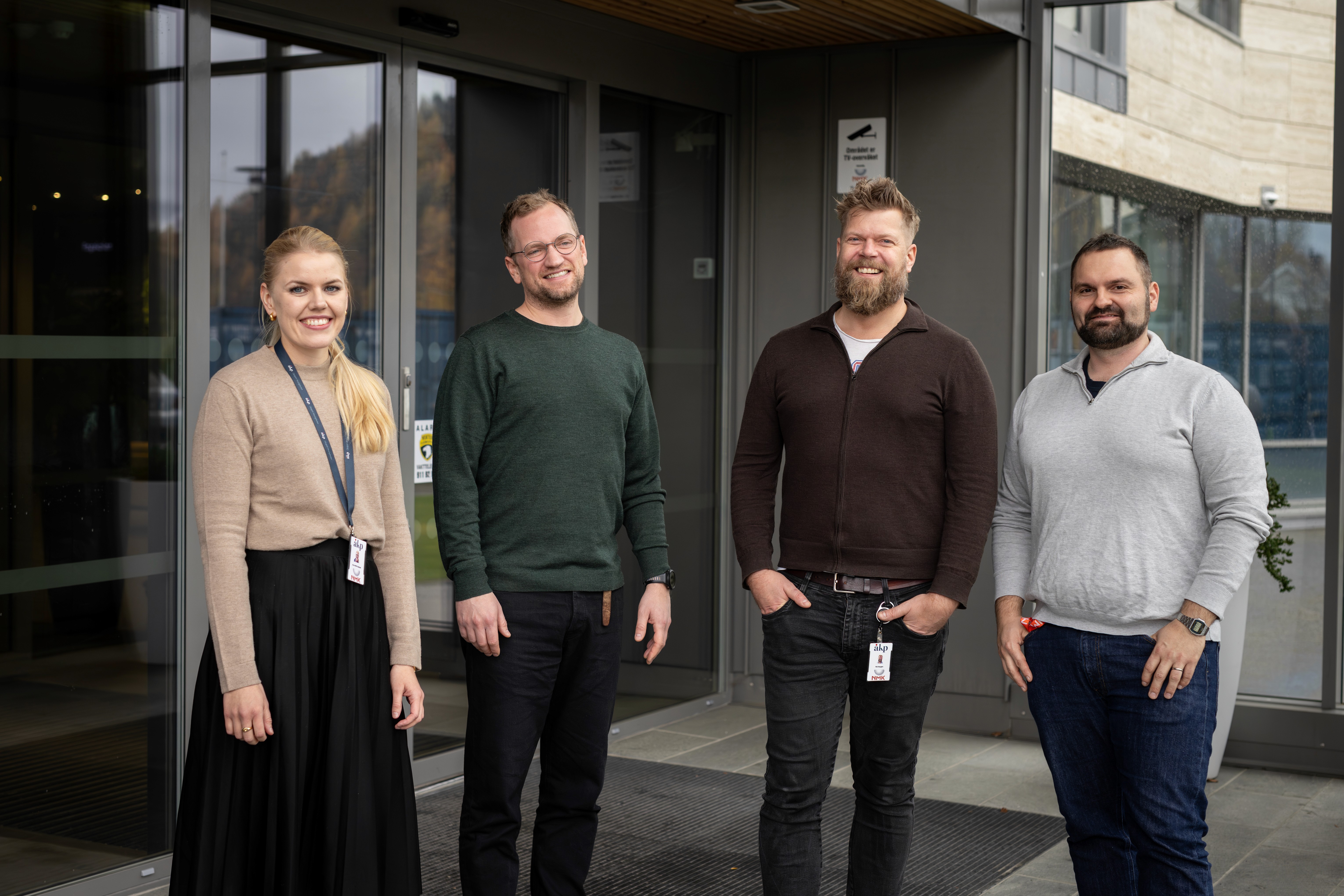 A group of four individuals standing outdoors in front of a building, smiling and dressed casually.