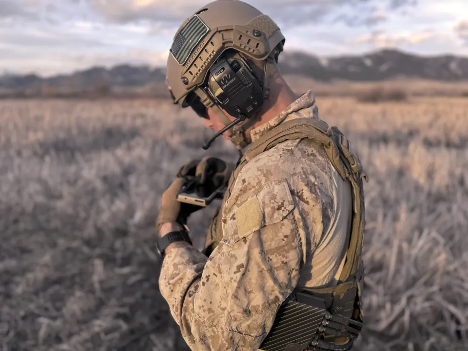 Marine in a helmet and headset checking a handheld device in a frosted field