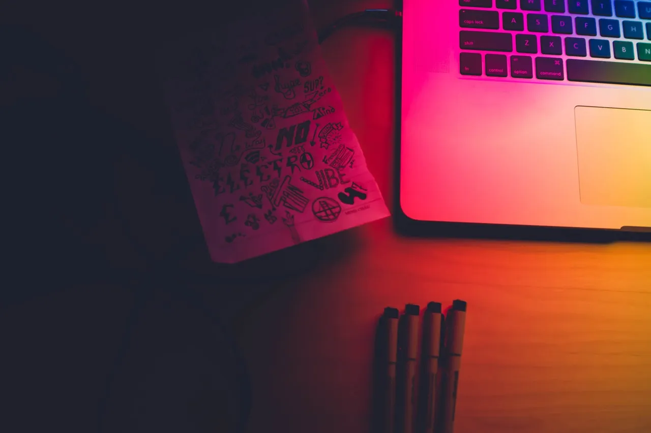 Creative workspace scene with a glowing laptop, sketchbook filled with doodles, and four markers on a wooden desk under moody red and orange lighting