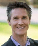 Smiling individual in a suit, outdoors with a blurred background of grass and trees.