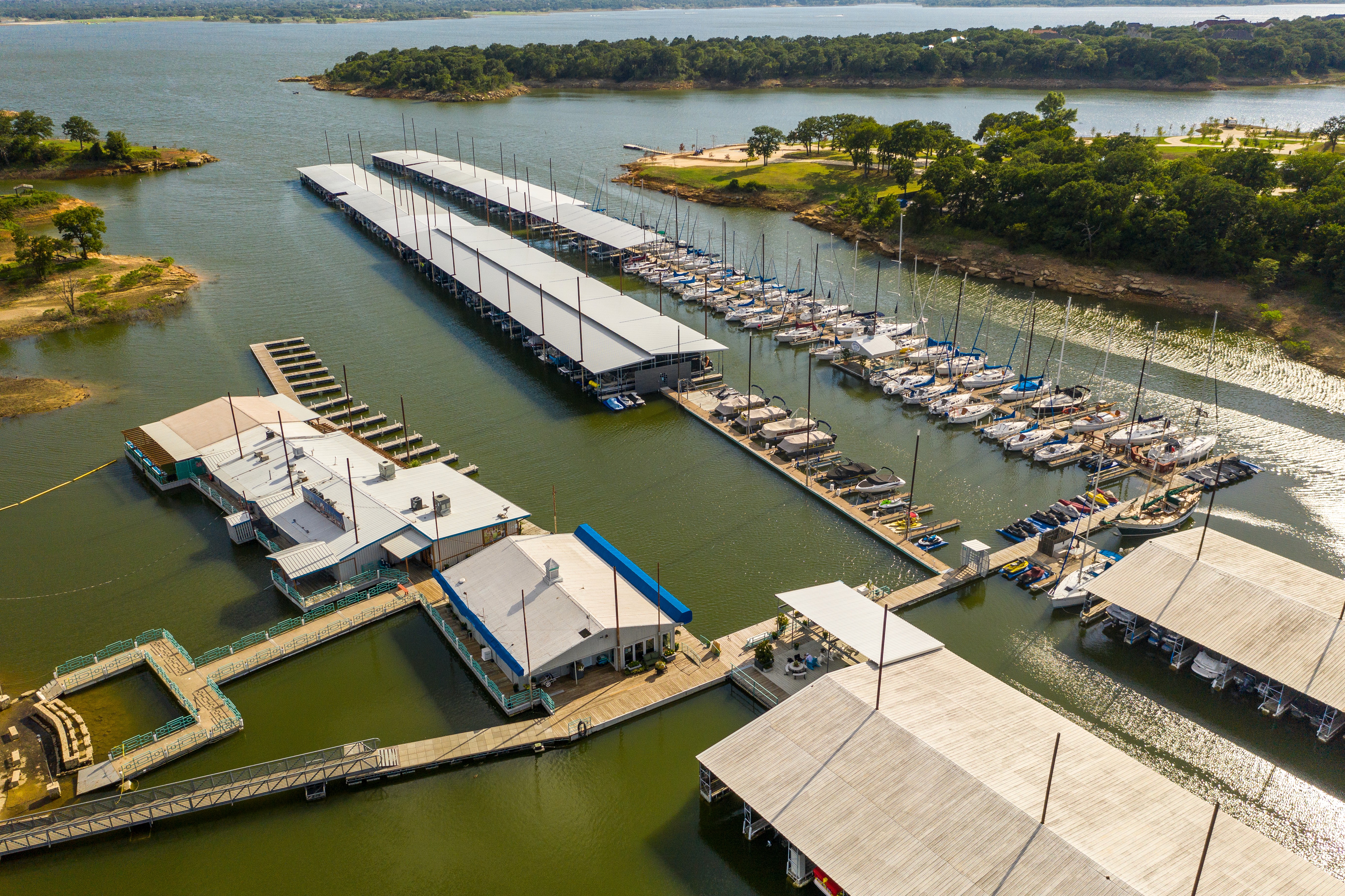Aerial view of a marina featuring multiple docks with numerous boats anchored, surrounded by calm lake waters and lush green trees, under a clear blue sky.
