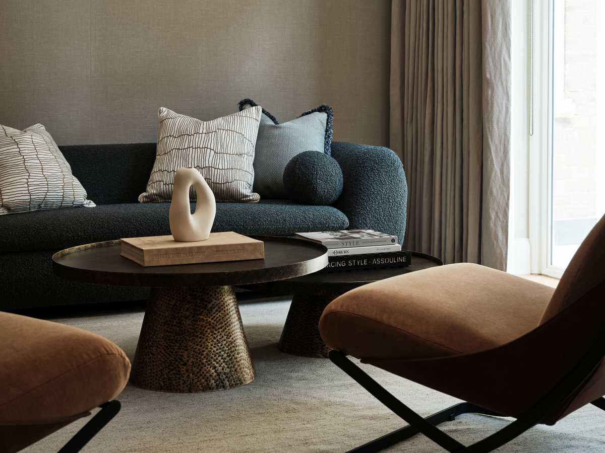 A cozy, high-end office lounge area featuring a deep charcoal boucle sofa with accent cushions, a textured bronze pedestal coffee table, and an ergonomic tan velvet armchair designed for long-term comfort.