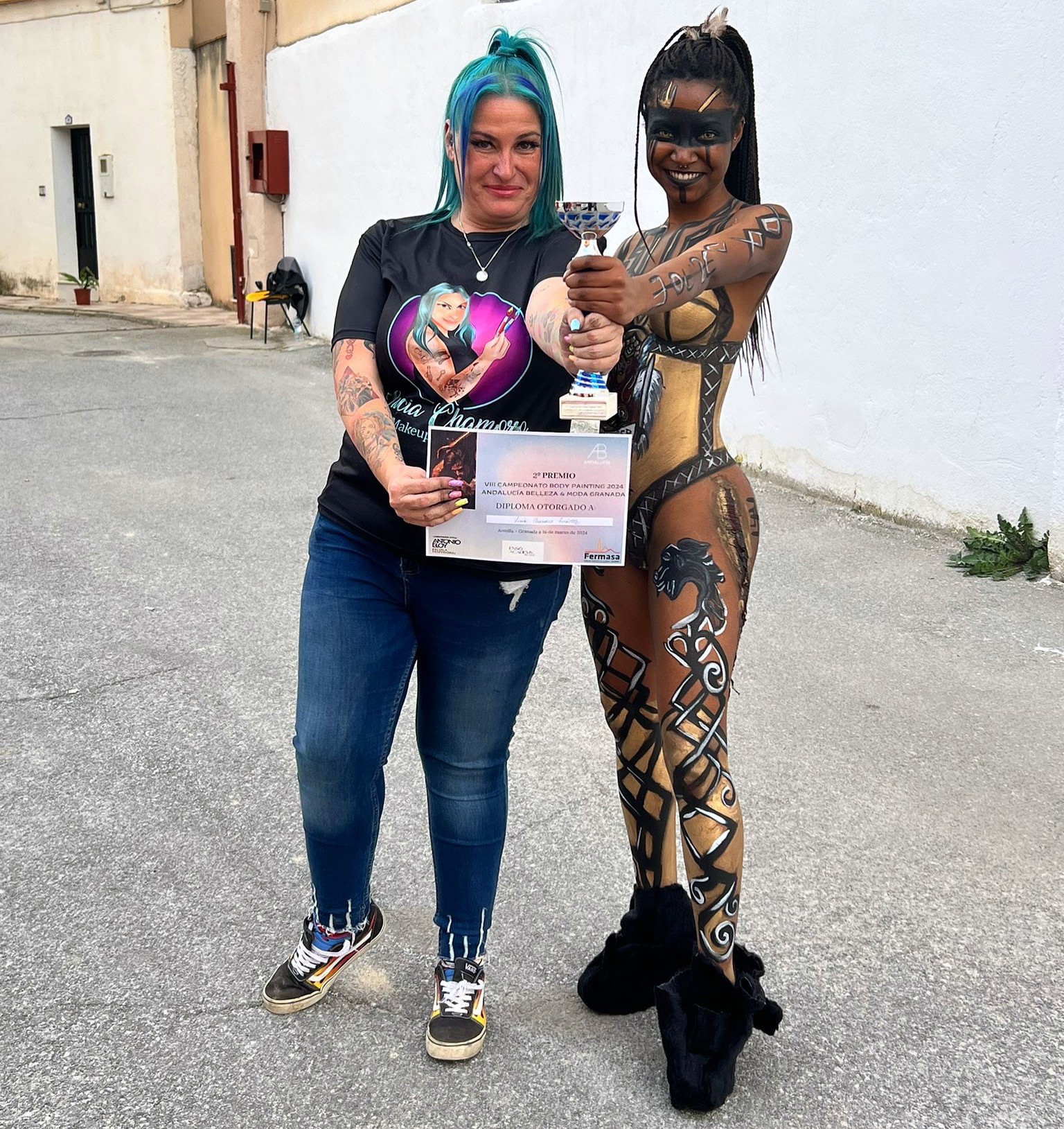 A young person with curly hair smiles at the camera while sitting in front of a laptop against a dark background.