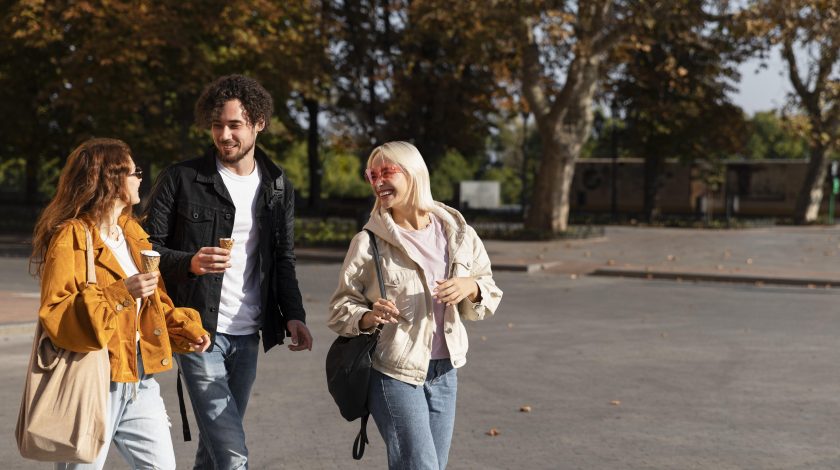 A group of three people dressed in casual autumn attire are joyfully walking on a sunlit street, carrying cups and bags, with autumn trees in the background.