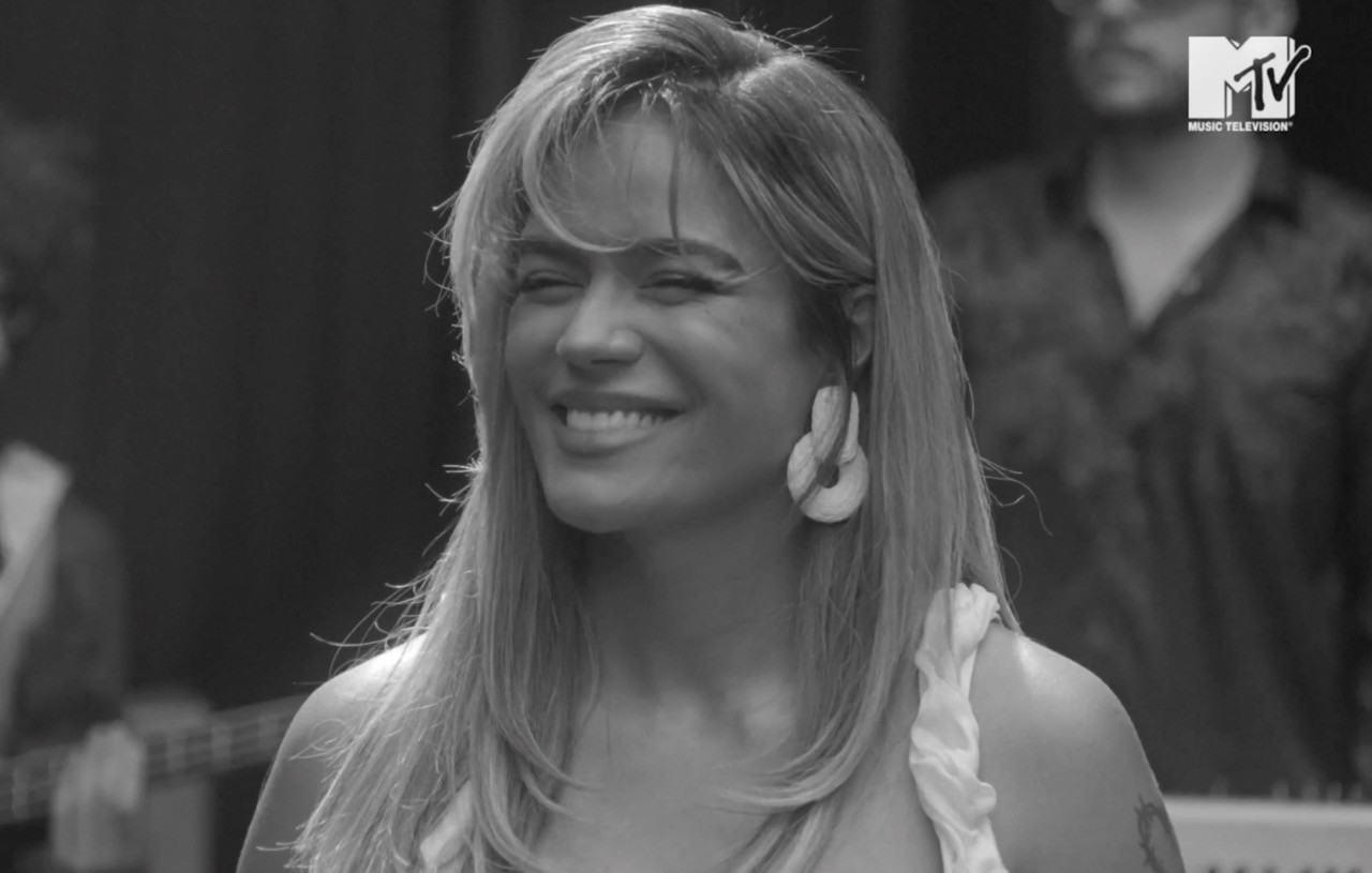 Black and white image of a Karol G smiling joyfully with closed eyes and long hair, wearing earrings and a sleeveless top. An MTV logo is in the corner.