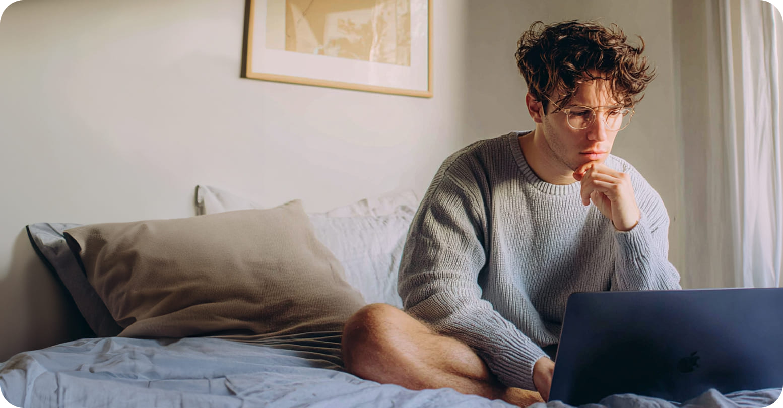 A young adult sitting on his bed looking at his laptop
