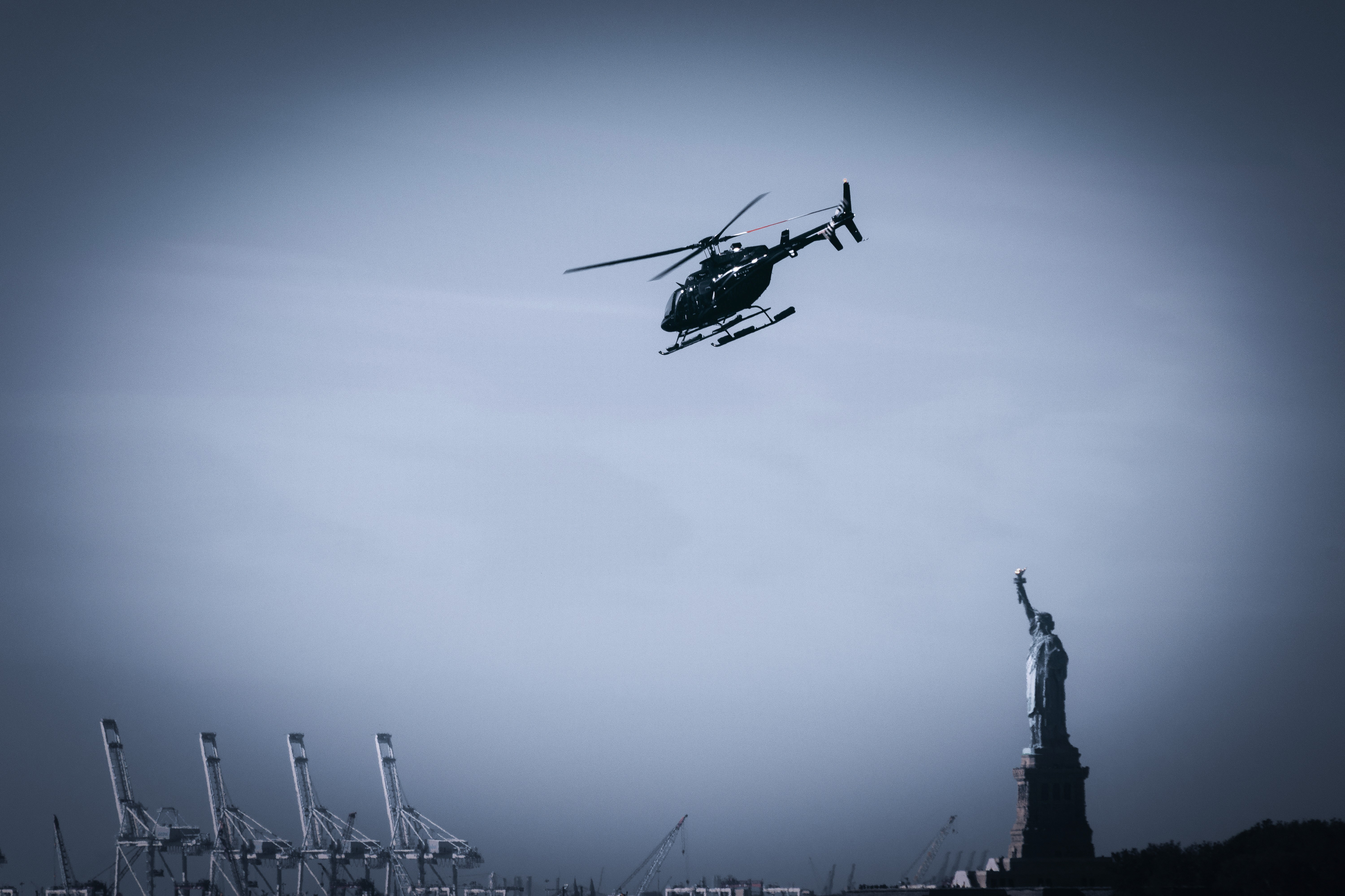 Schwarzer Hubschrauber fliegt an einem bewölkten Tag über die Freiheitsstatue, im Hintergrund sind Industriekräne zu sehen.