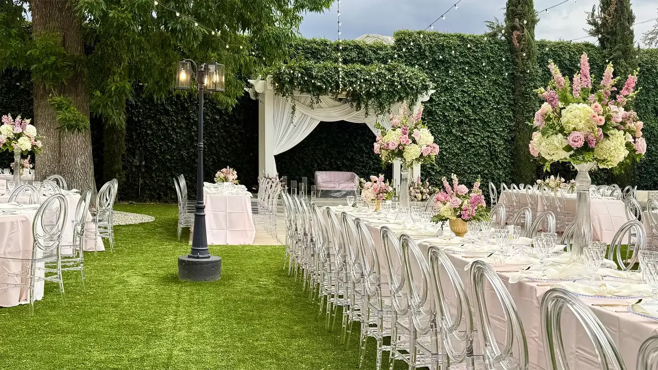 Long pink-linen tables with tall floral centerpieces and clear acrylic chairs on a lawn, next to an ivy wall and a draped arbor.