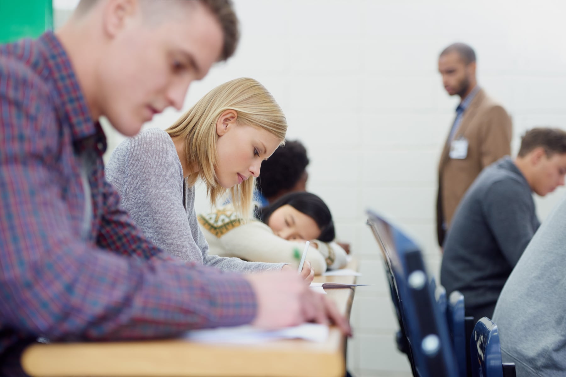 A classroom of students taking an exam, with a student in the centre focused on writing while an invigilator walks behind them.